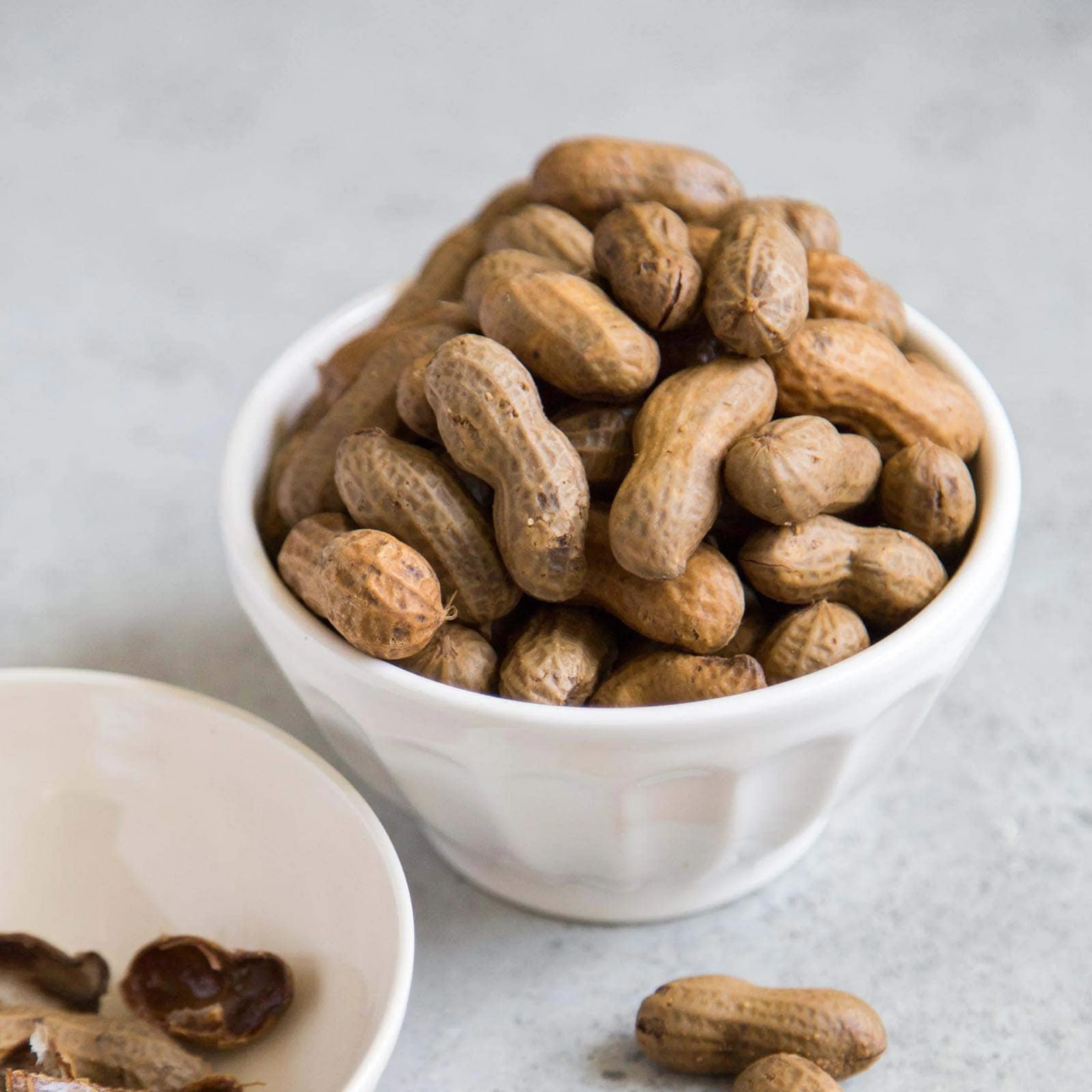 A white bowl piled with whole, unshelled peanuts sits on a pale stone surface, with a few loose peanuts and a partial dish nearby; the scene feels calm and rustic.