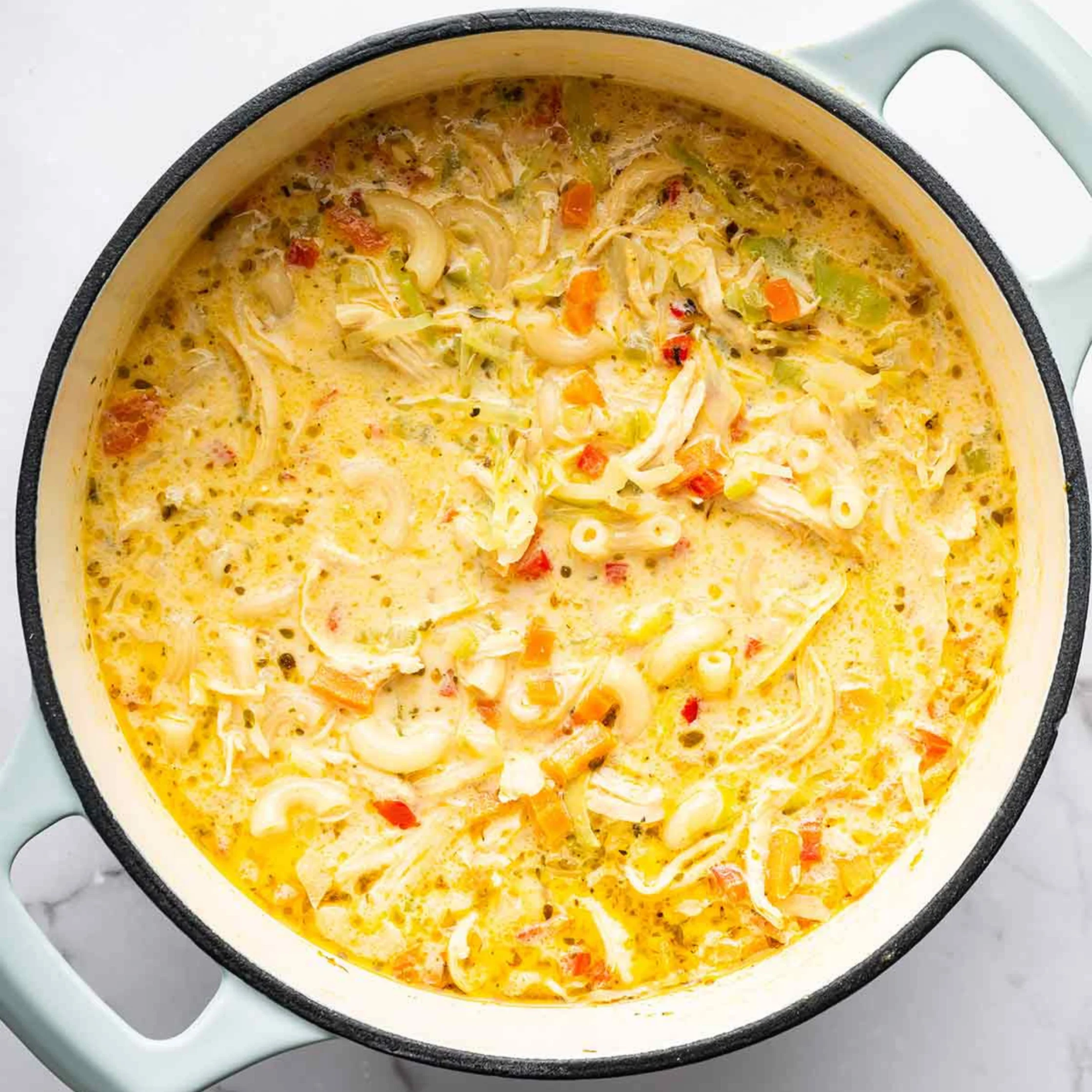 Overhead view of a creamy chicken noodle soup with shredded chicken, pasta, carrots, and herbs in a pale blue pot; warm, comforting meal.