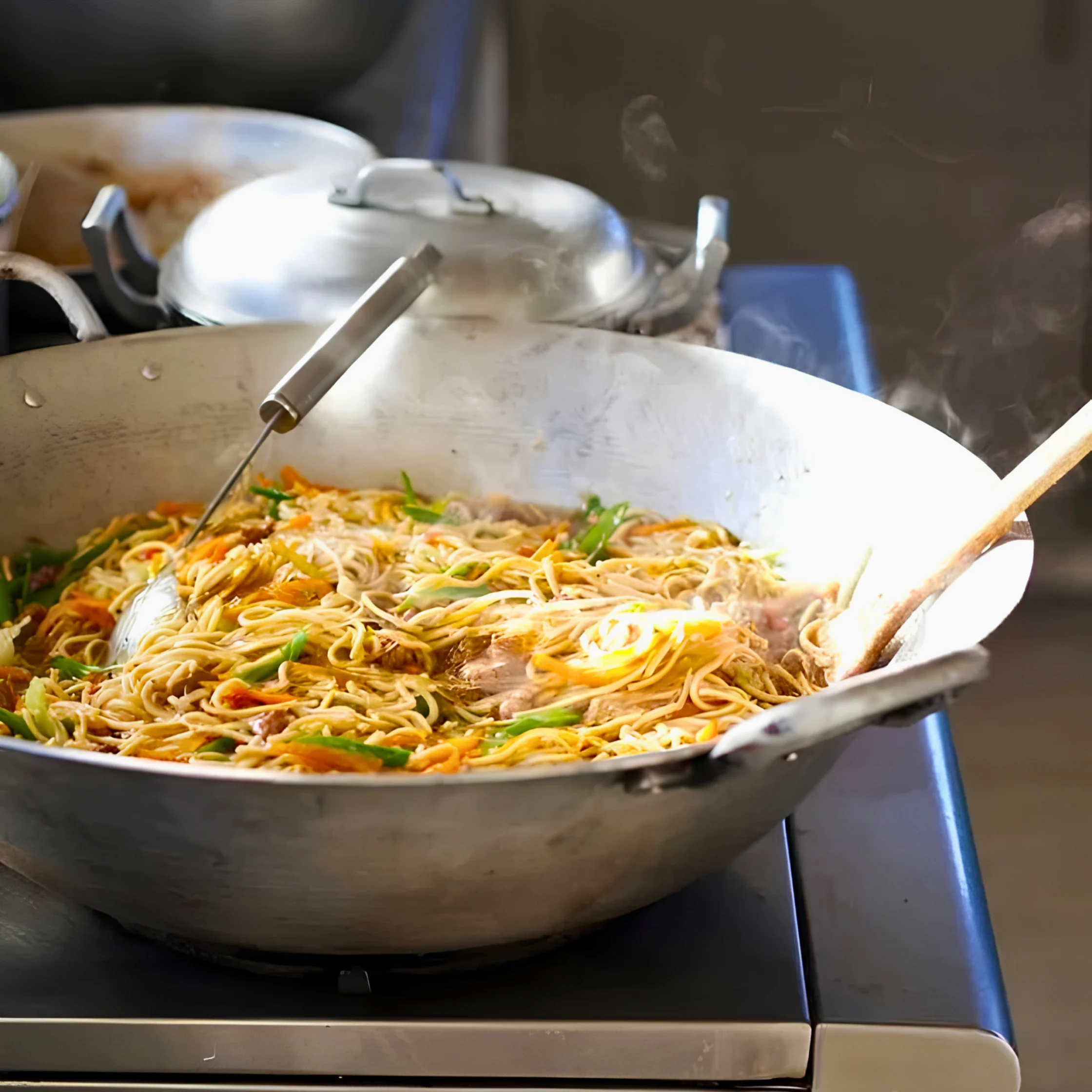 A large metal wok of steaming stir-fried noodles with vegetables and herbs sits on a stovetop, evoking the warmth and bustle of a kitchen.