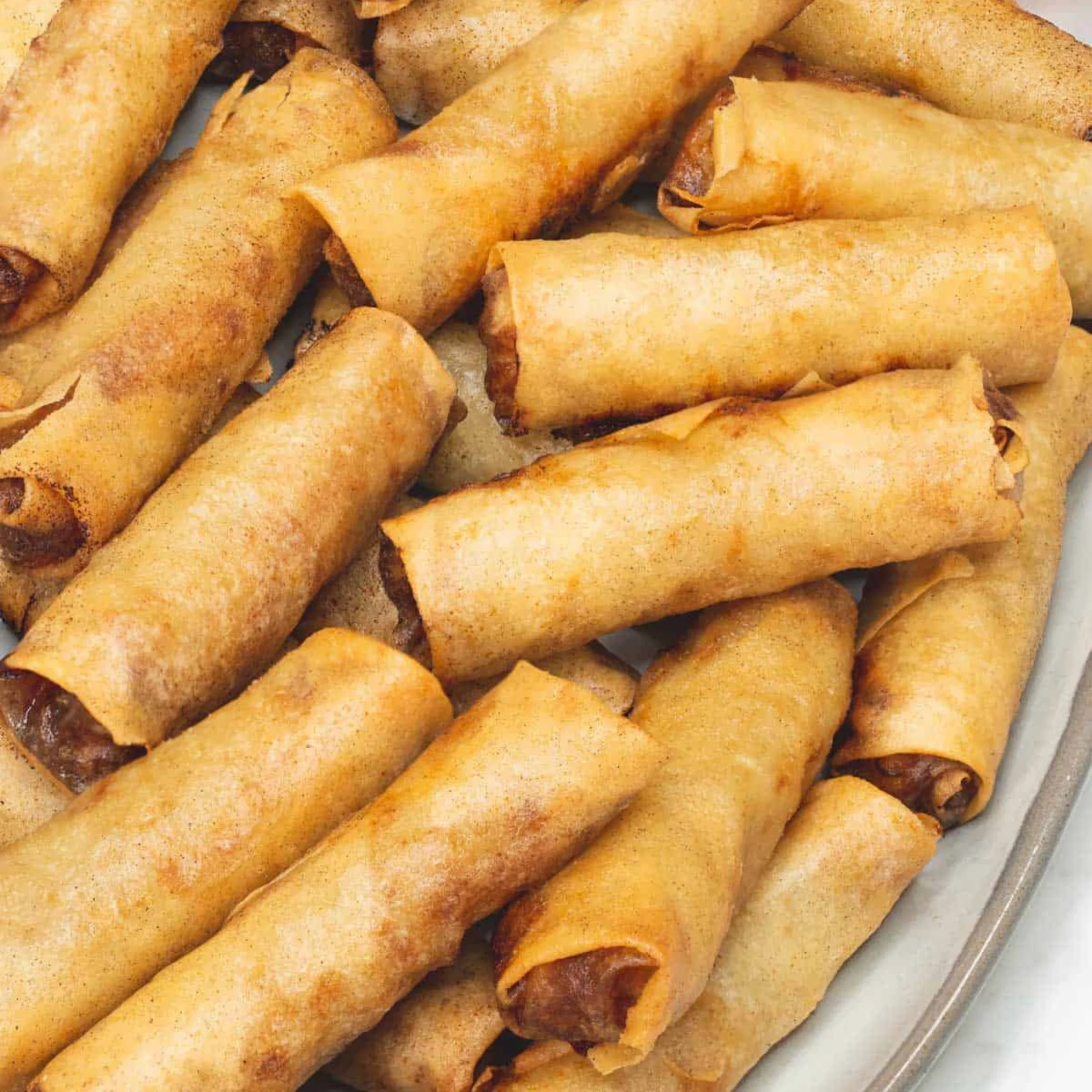 A plate piled with golden-brown, crispy spring rolls, their flaky wrappers showing hints of savory filling; the close-up feels warm and appetizing.