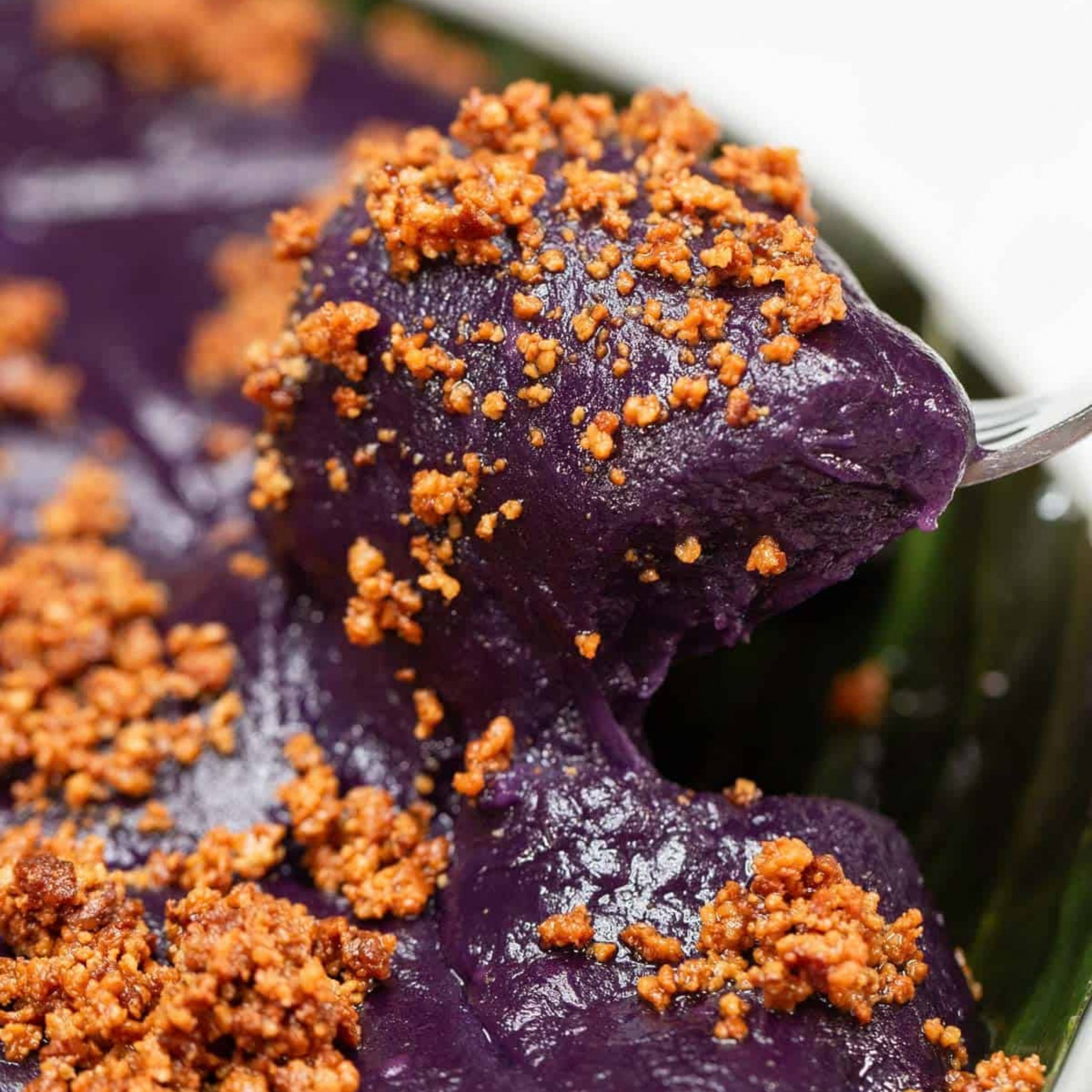 Close-up of glossy purple mashed sweet potatoes topped with golden-brown crunchy crumbs, with a spoon lifting a rich scoop; the dish looks hearty and inviting.