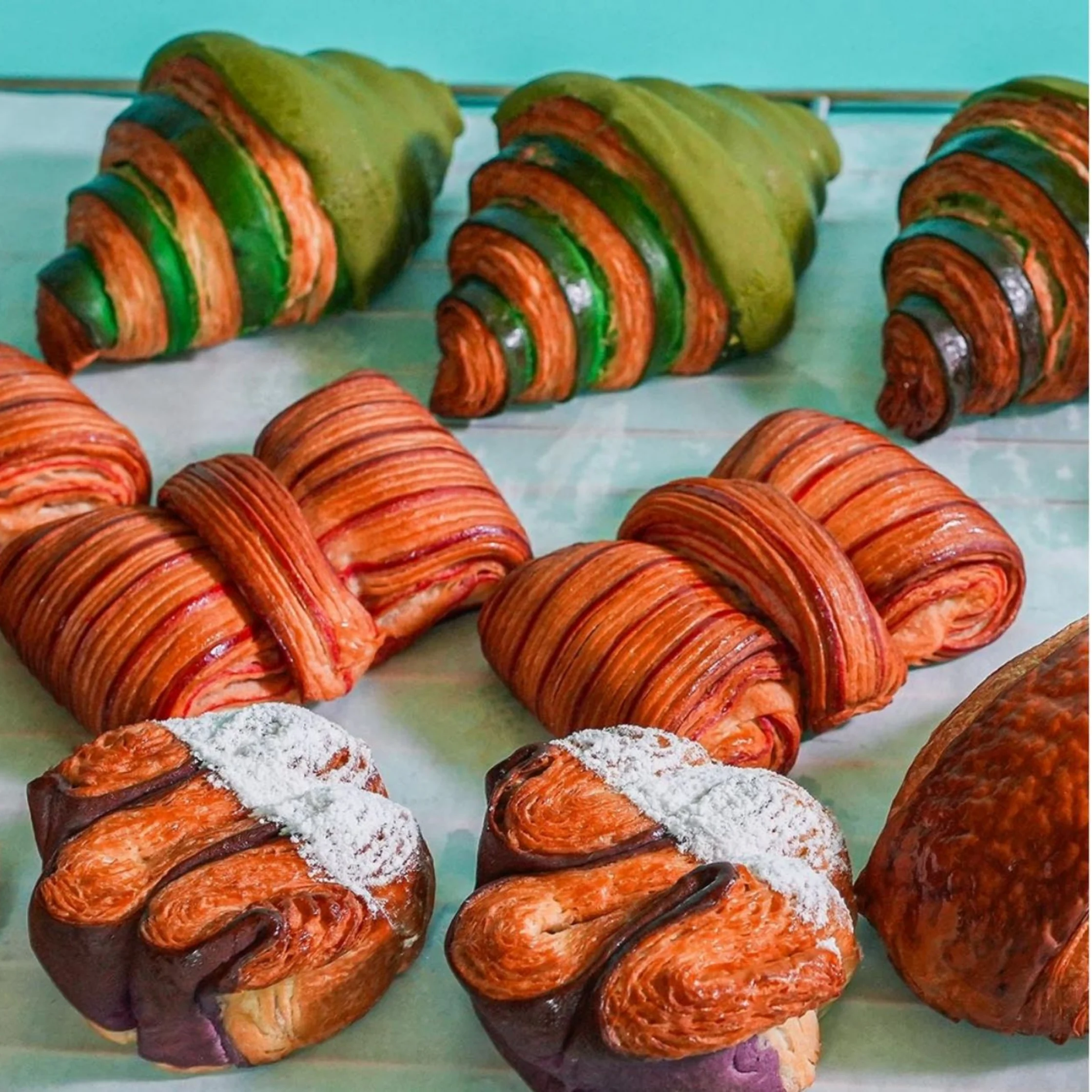 Colorful croissants and laminated pastries fill a bakery tray, including green-and-orange rolls and powdered-sugar twists, creating a vivid, inviting display.