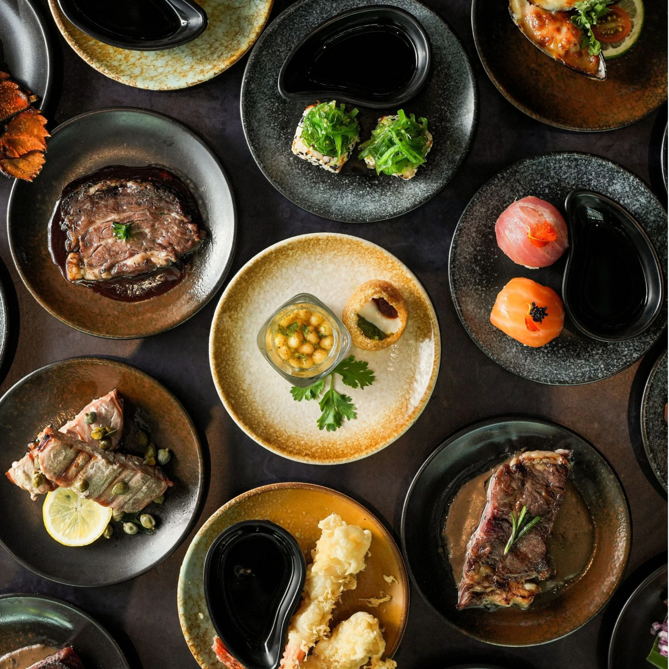 Overhead spread of gourmet dishes on dark plates: sushi, grilled meat, seafood, greens, and sauces, arranged artfully for an elegant dining mood.