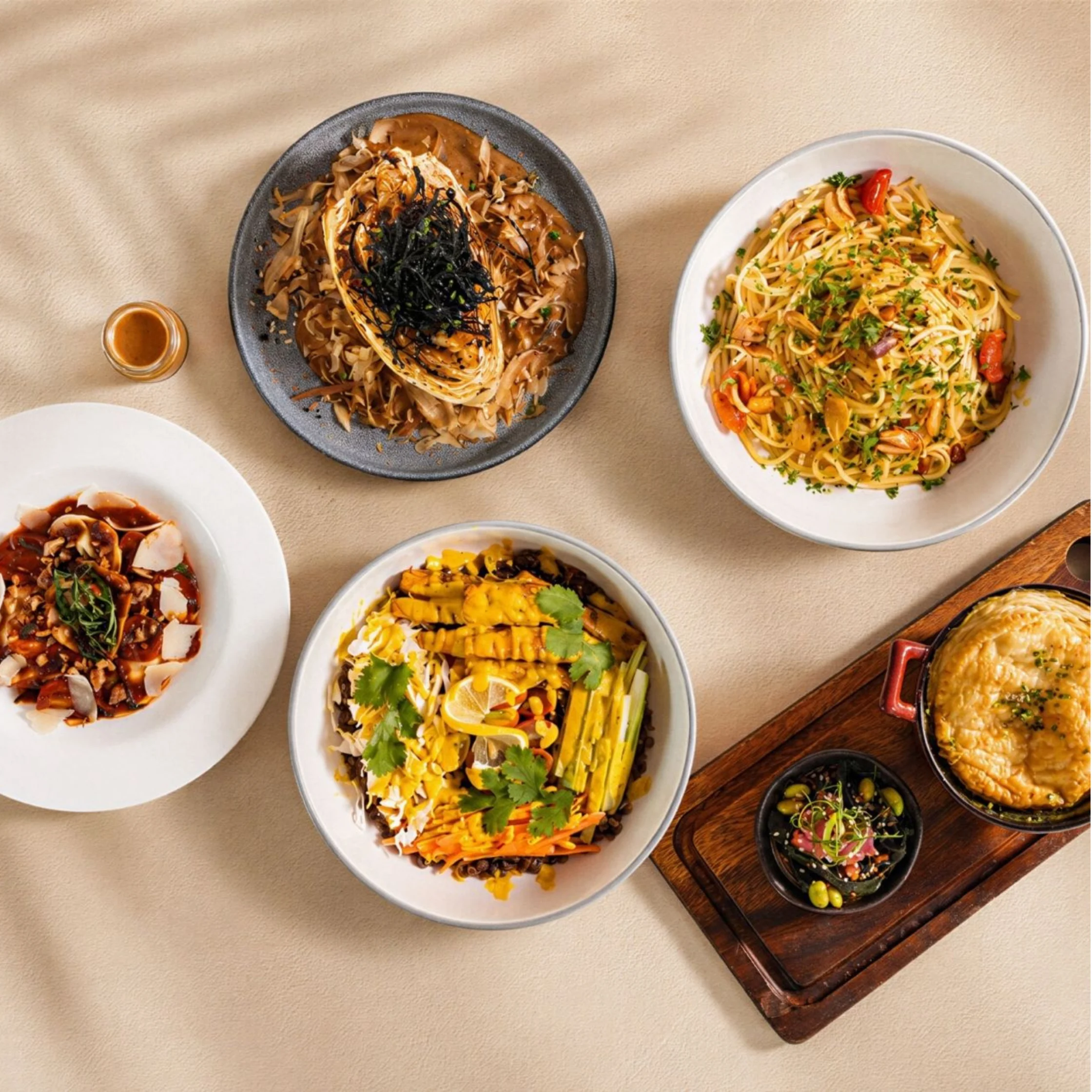 Overhead spread of colorful Asian dishes—noodles, rice, curry, and stew—on ceramic plates and a wooden board, arranged invitingly on a beige table.