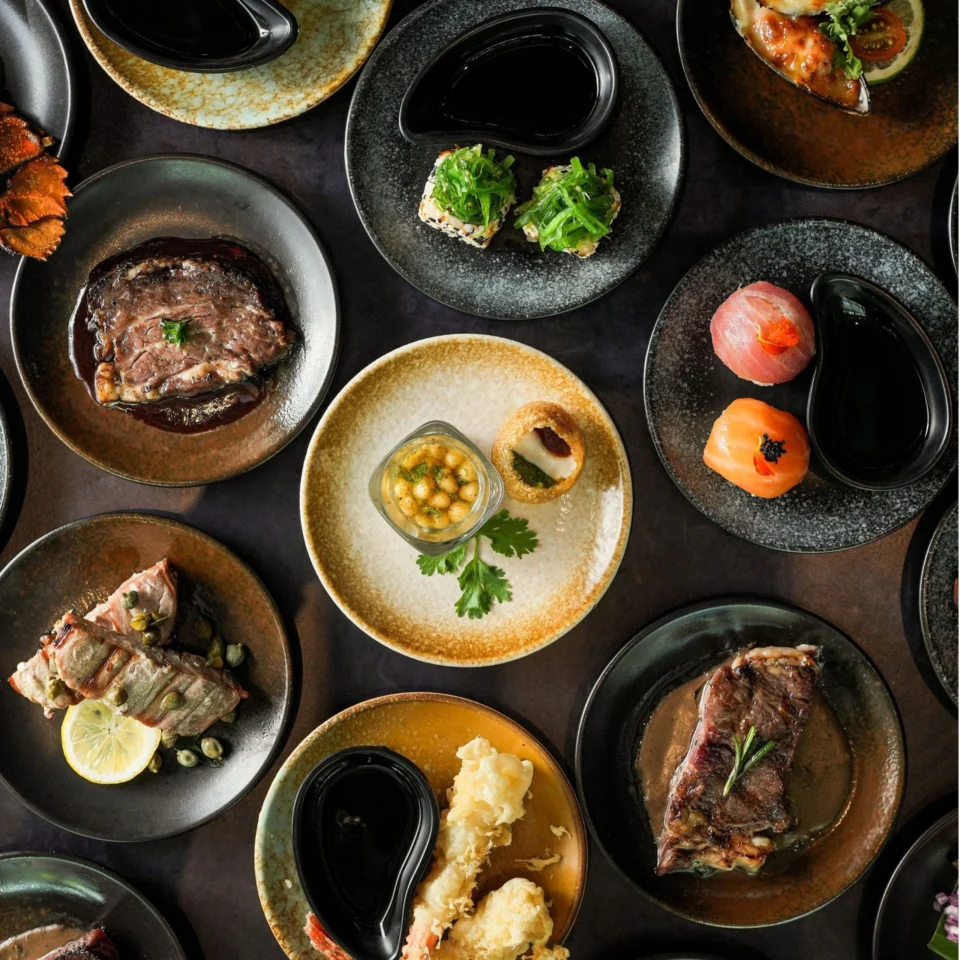 Overhead spread of gourmet dishes on dark plates: sushi, grilled meat, seafood, greens, and sauces, arranged artfully for an elegant dining mood.