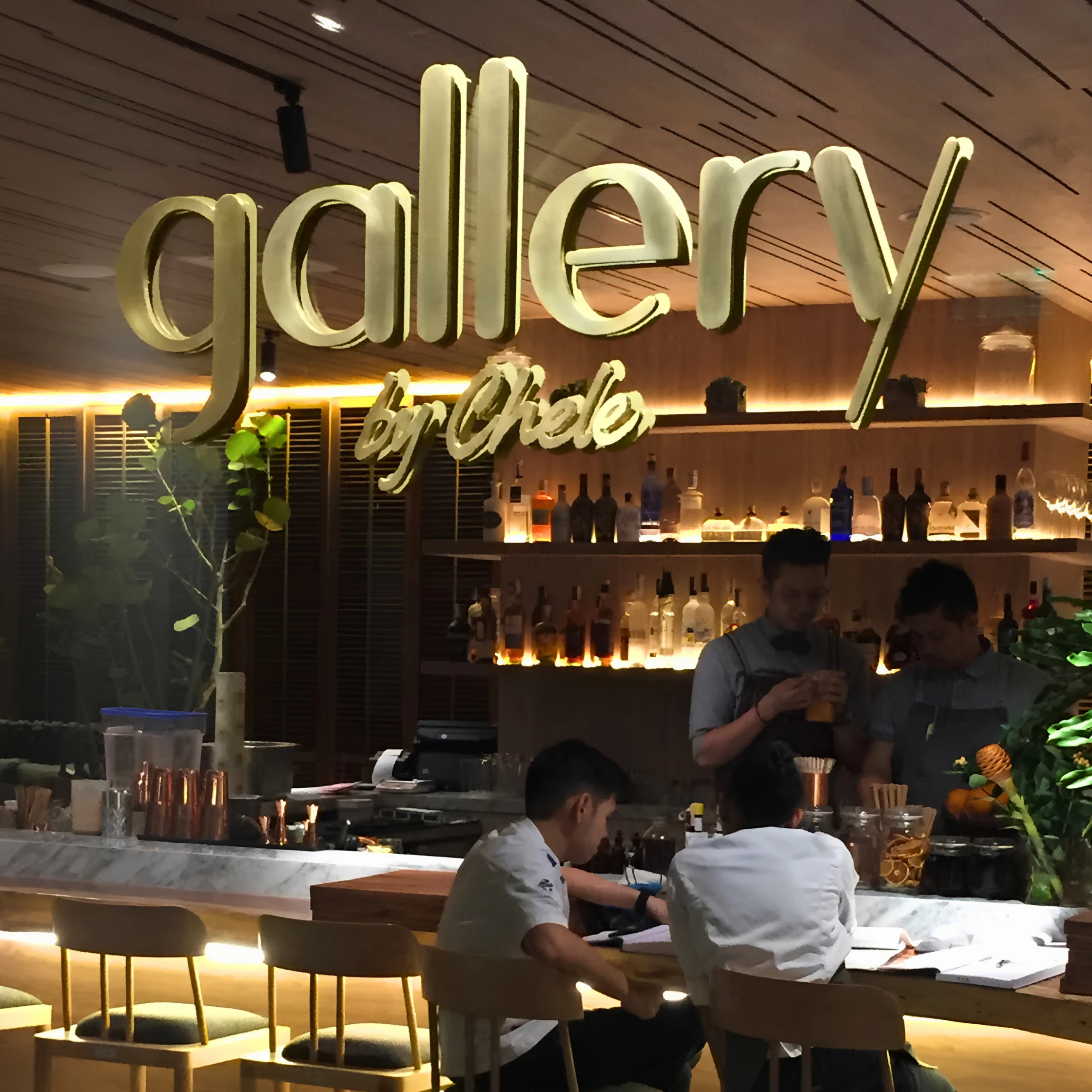 A warmly lit modern bar with large gold “gallery by Chele” lettering, glowing bottle shelves, and staff serving diners seated at the counter.