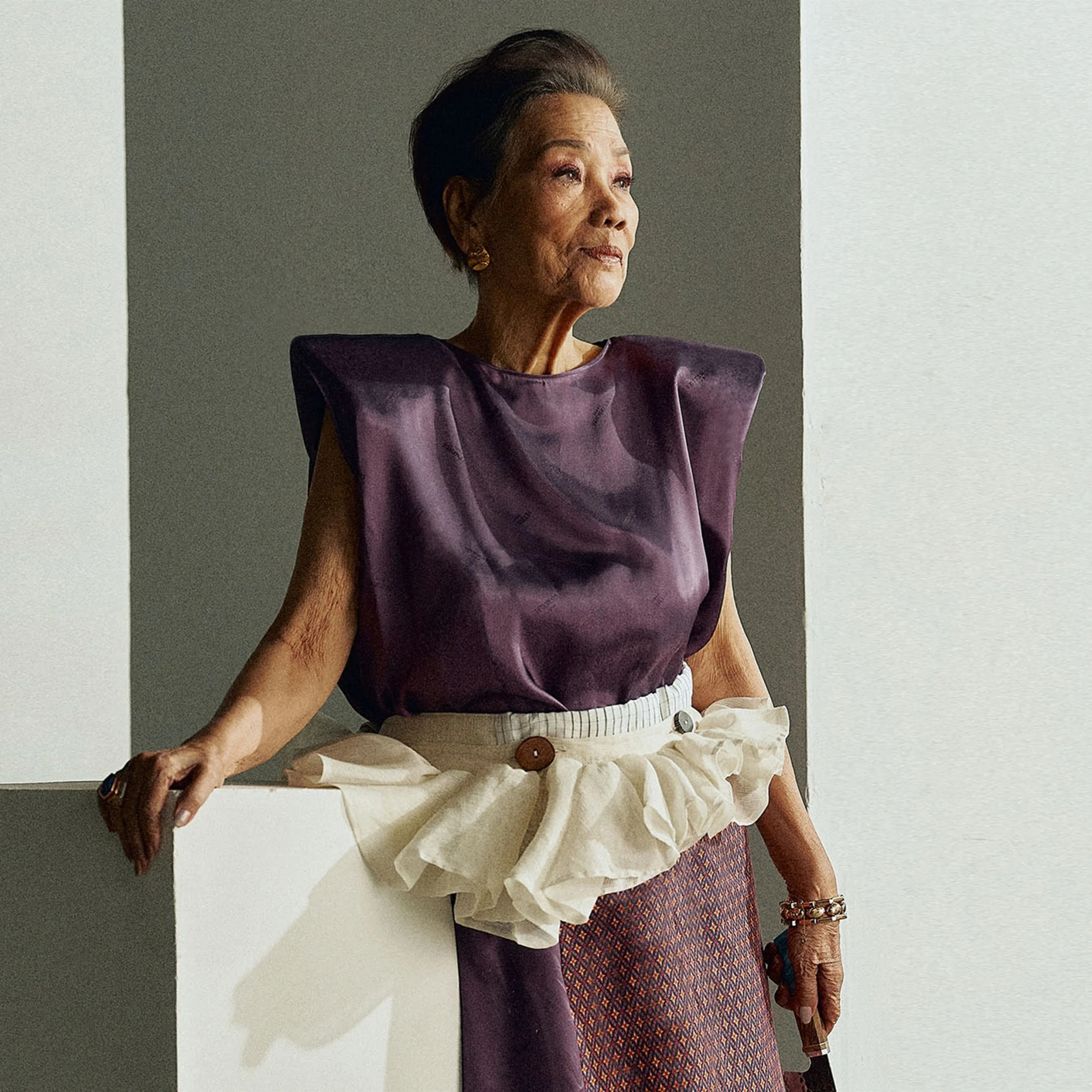 An older woman in a purple sculptural top and layered cream skirt poses beside a wall, gazing into the light with quiet confidence and poise.