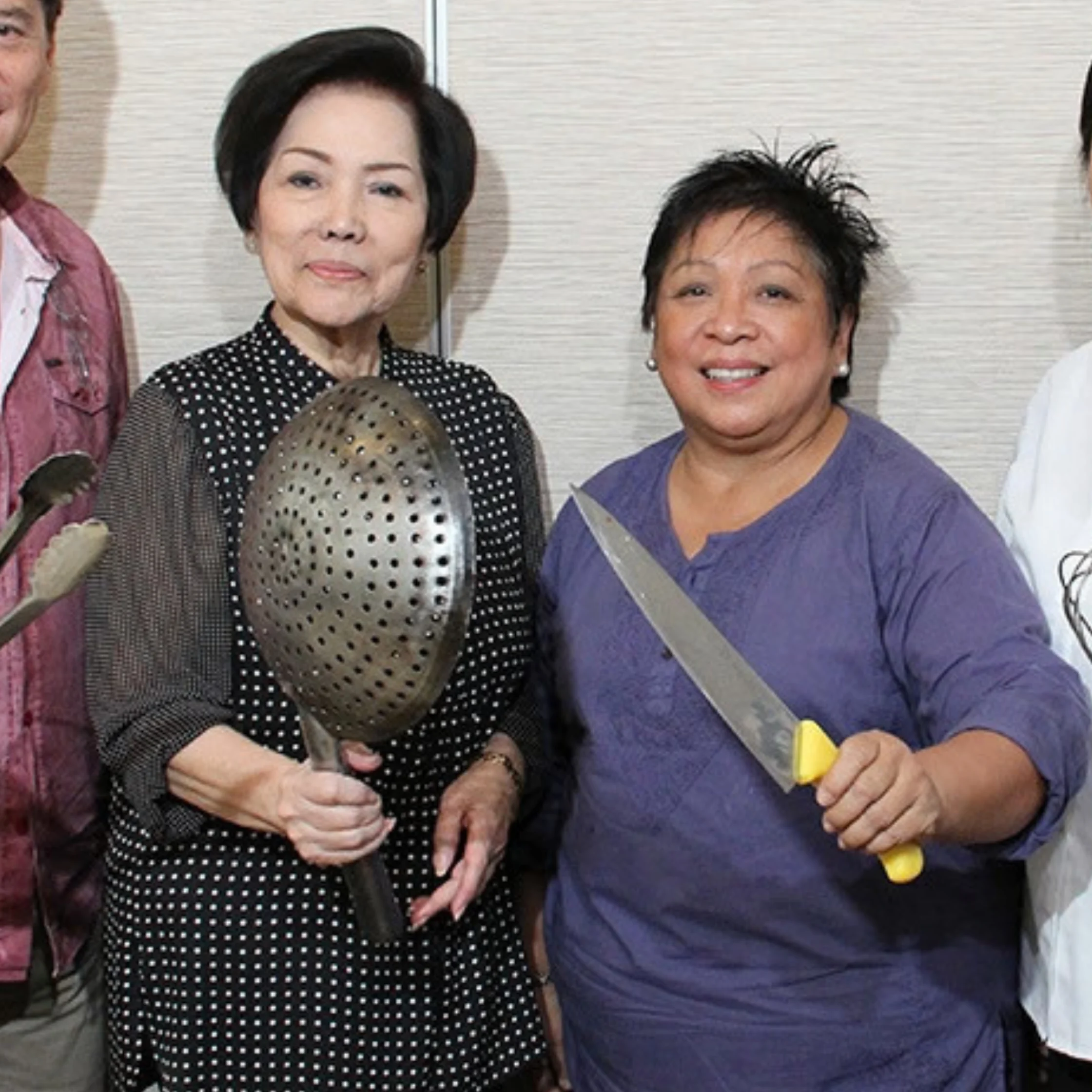 Two smiling women pose indoors with oversized kitchen utensils: one holds a perforated skimmer, the other a large knife. The playful photo has a warm, lighthearted tone.