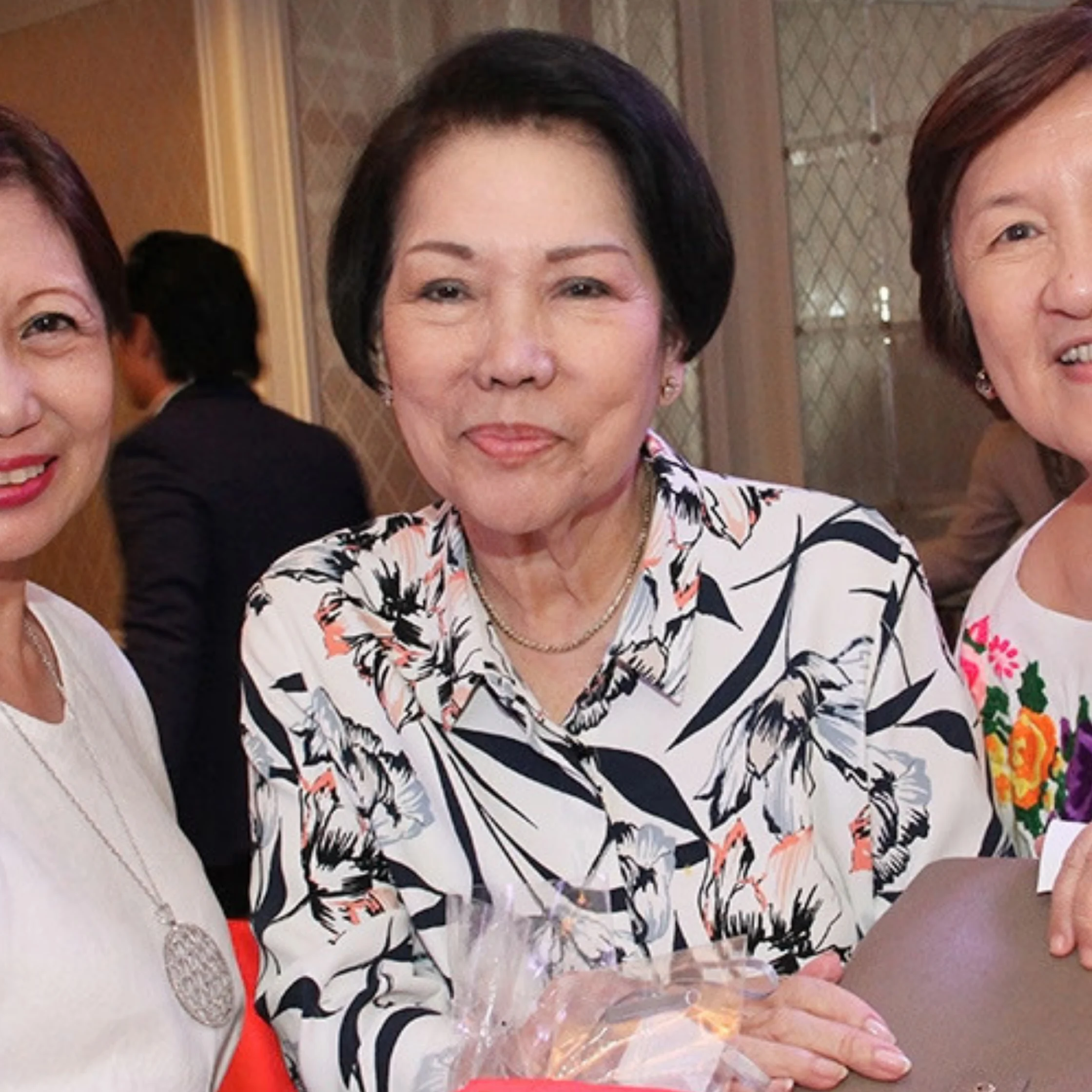Three smiling older women pose indoors, with a woman in a floral blouse at center holding a wrapped gift; the candid gathering feels warm and celebratory.