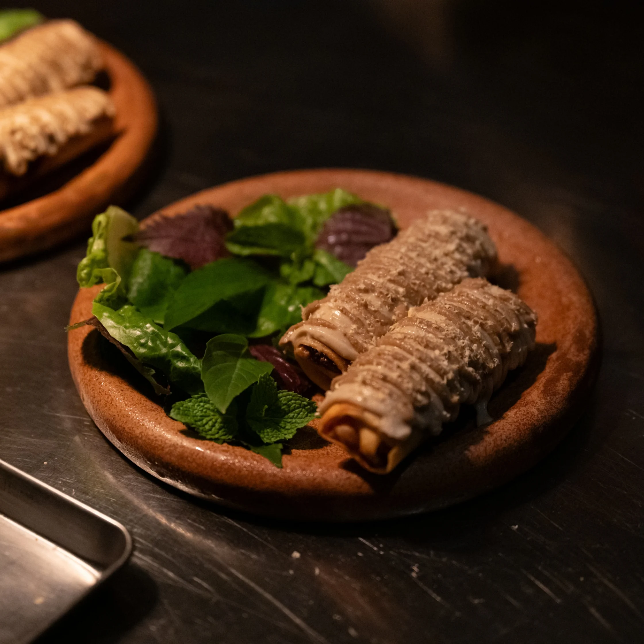 Two crisp, rolled savory pastries sit beside fresh leafy greens on a rustic brown plate; warm, low lighting gives the food a cozy, appetizing feel.