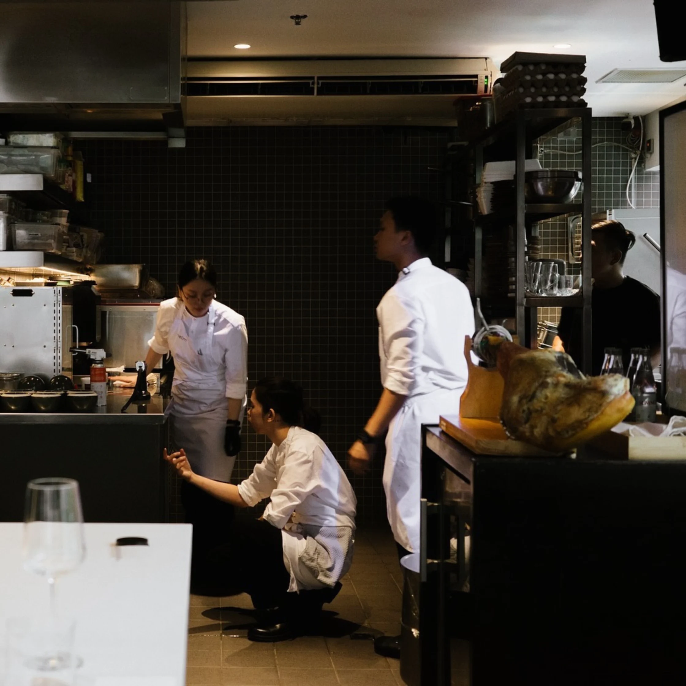 Chefs in white uniforms confer in a dim, busy restaurant kitchen beside a large cured ham; the focused scene feels intense and professional.
