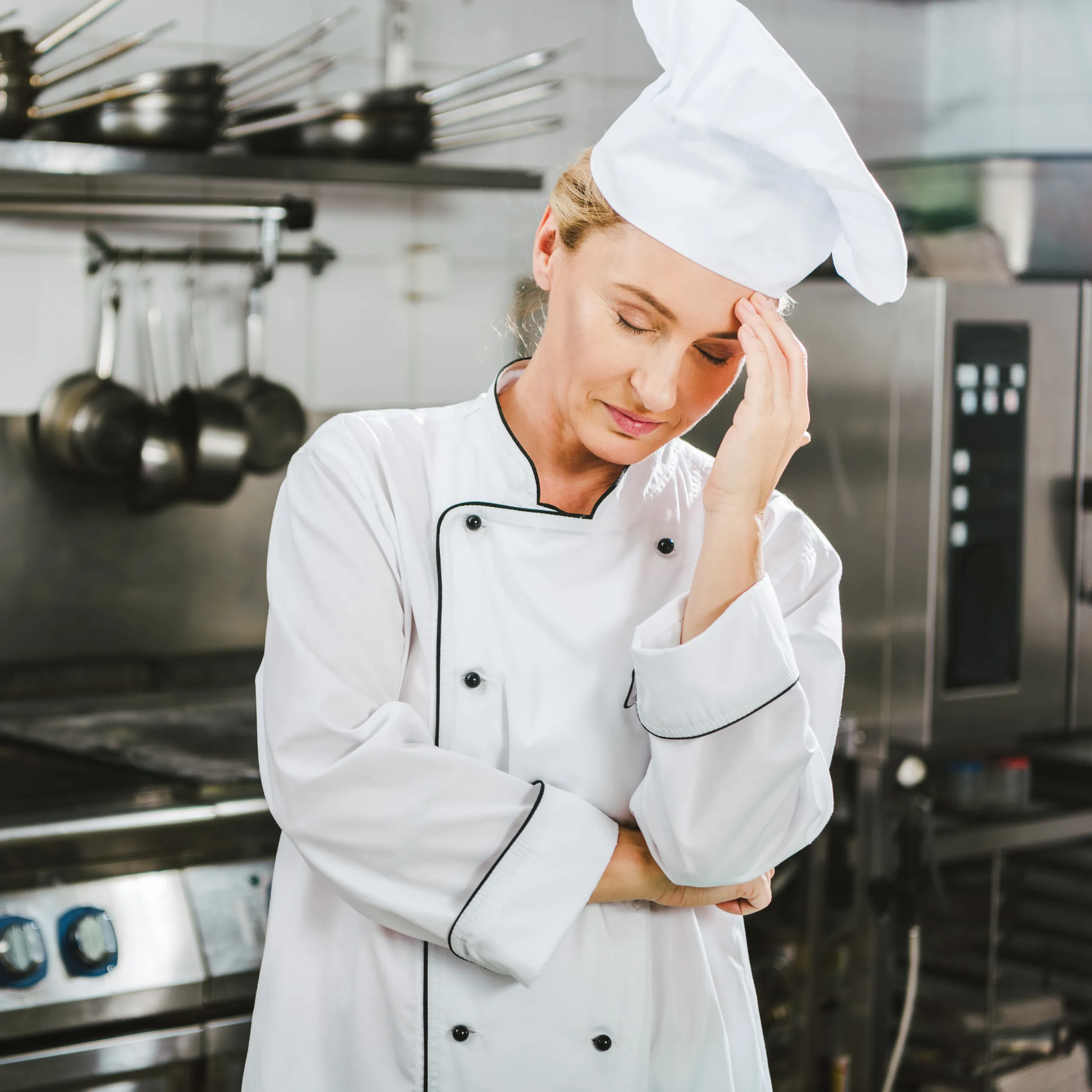 A tired woman chef in a white uniform and tall hat rests her hand on her forehead in a stainless-steel commercial kitchen, conveying fatigue.