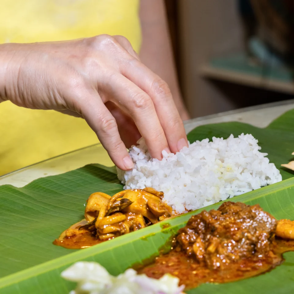 A hand gathers white rice beside spicy curry on a green banana leaf, showing a traditional South Asian meal eaten by hand in a casual setting.