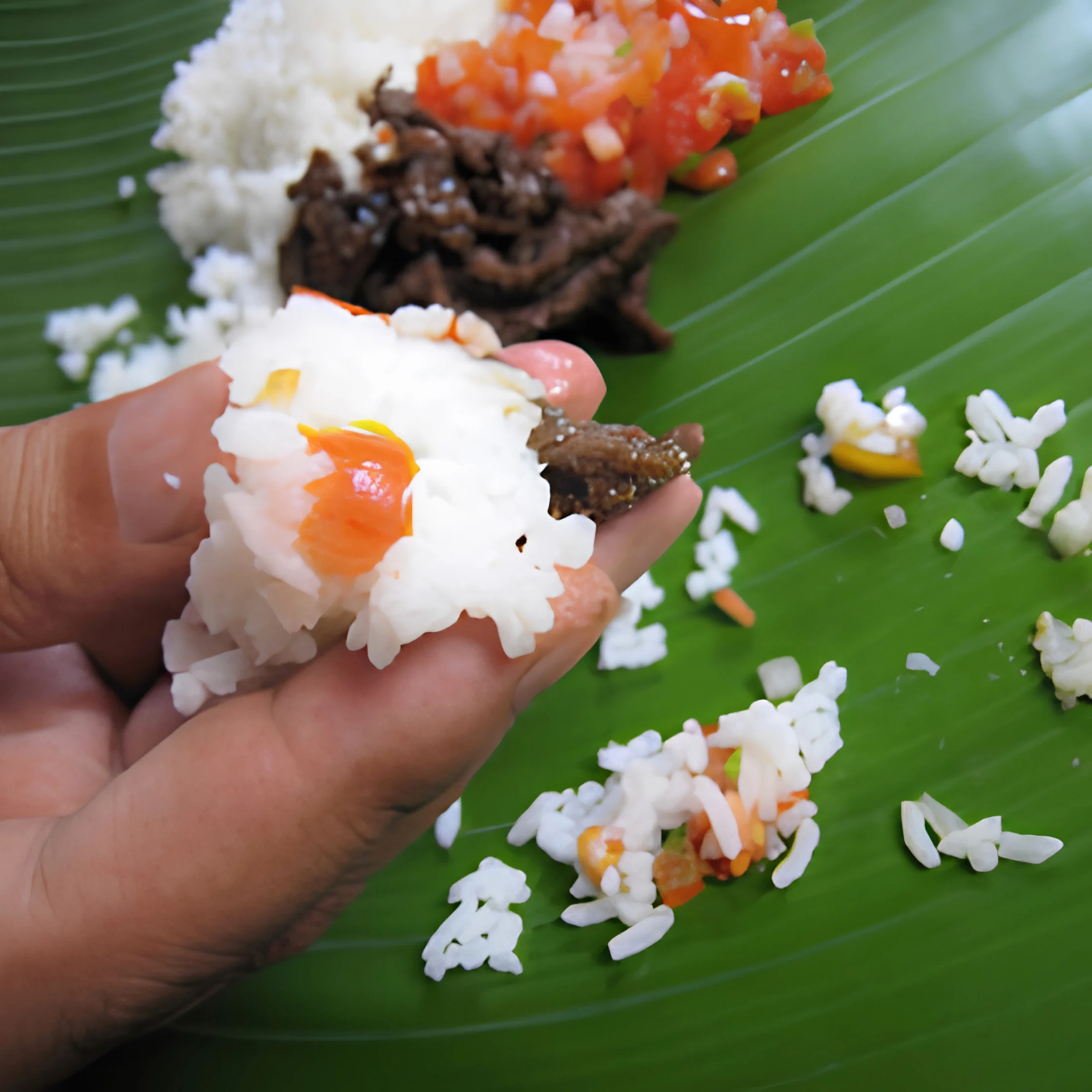 A hand holds white rice topped with tomato salsa and browned meat over a green banana leaf, with more food and scattered rice behind it.