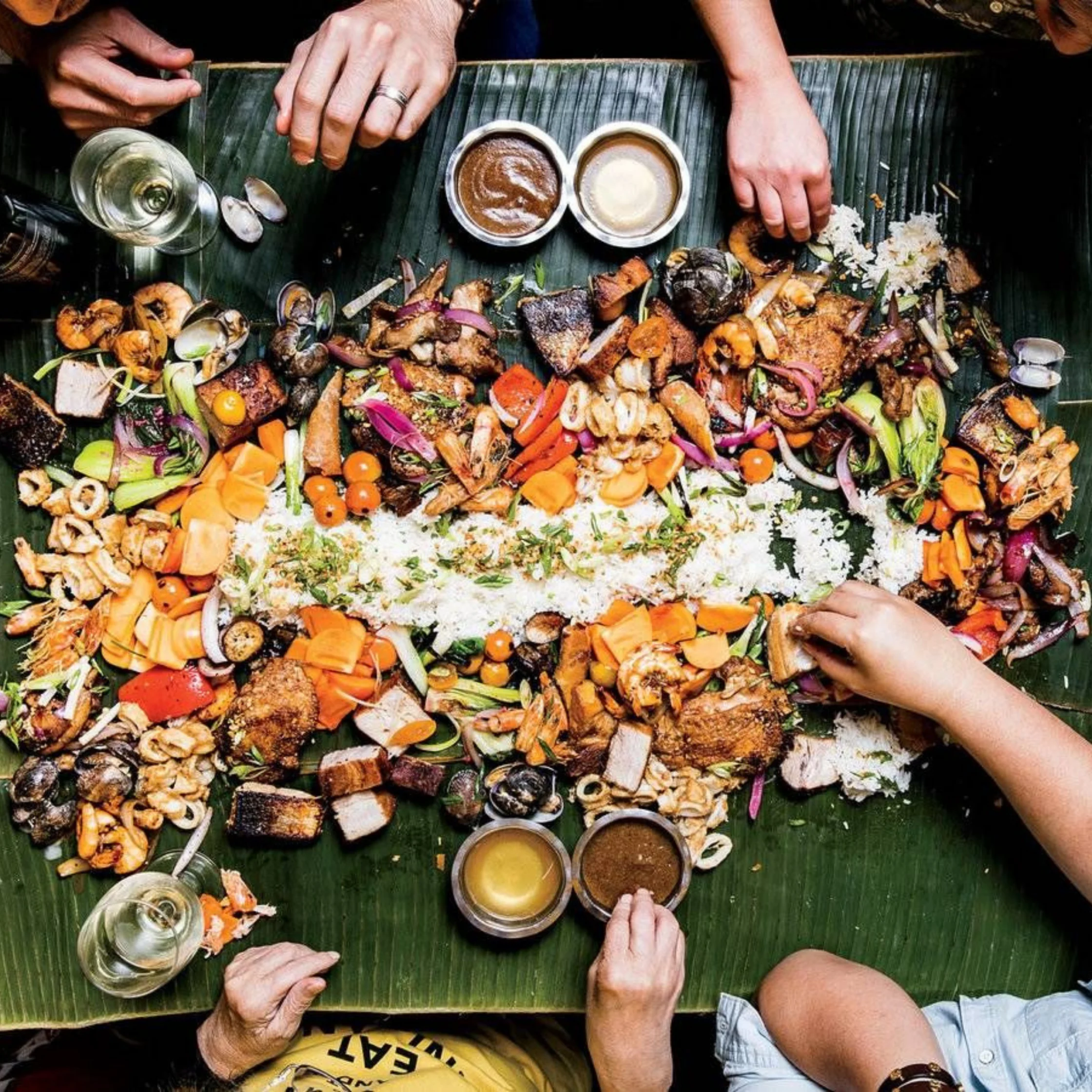 Overhead view of hands sharing a colorful seafood feast with grilled vegetables, rice, and dipping sauces on a banana leaf, creating a lively communal mood.