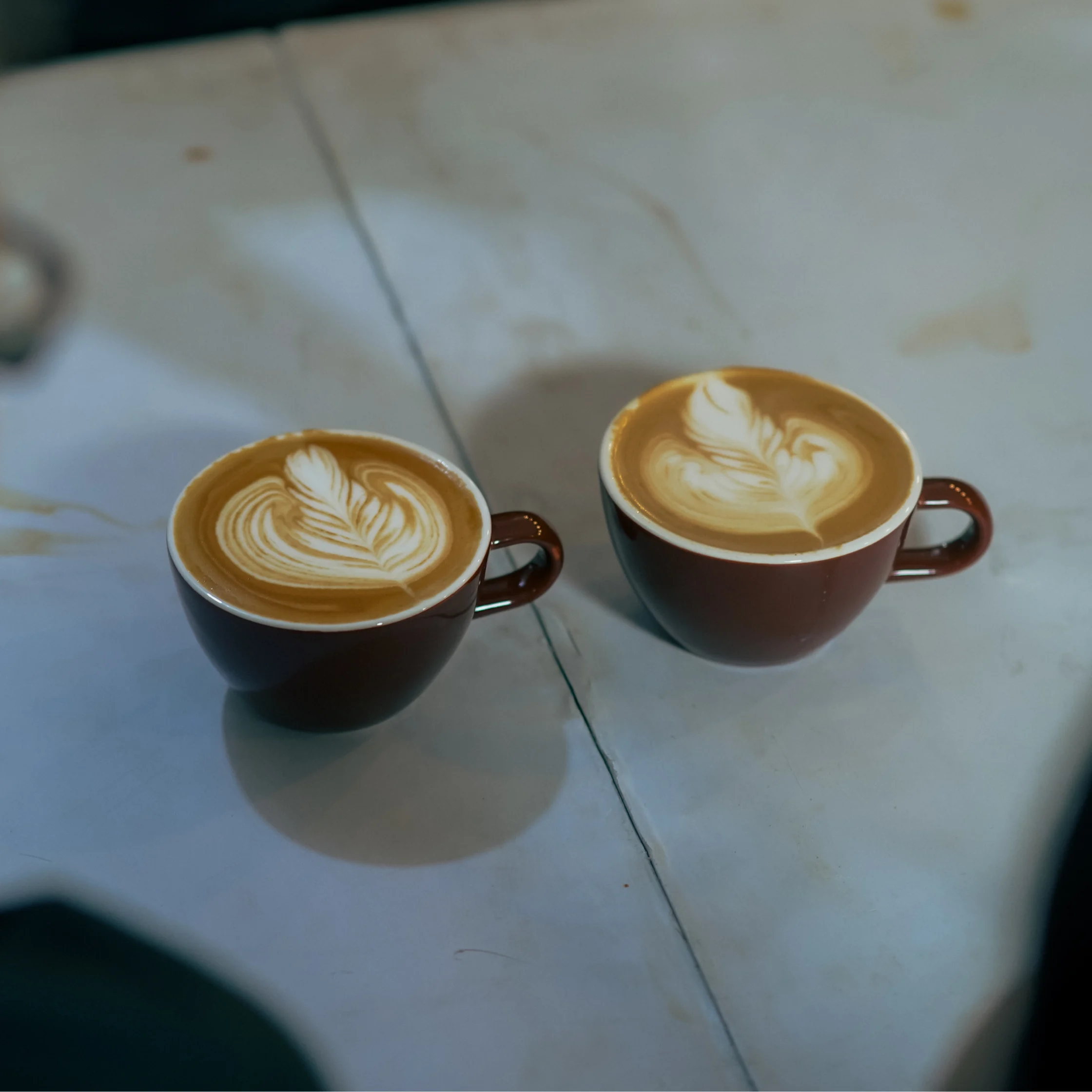 Two ceramic cups of latte sit on a pale stone table, each topped with intricate heart-shaped foam art; the warm coffee scene feels calm and inviting.
