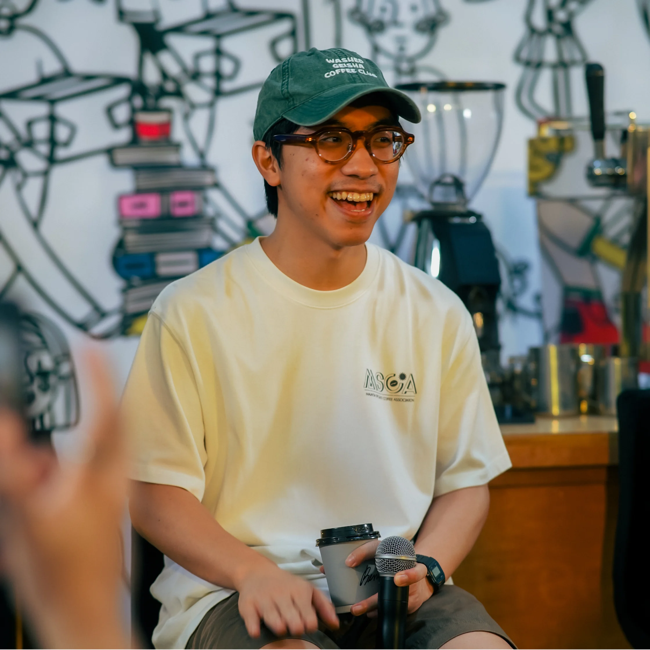 A smiling young man in a green cap, glasses, and cream T-shirt holds a microphone in a coffee shop, with brewing equipment behind him.