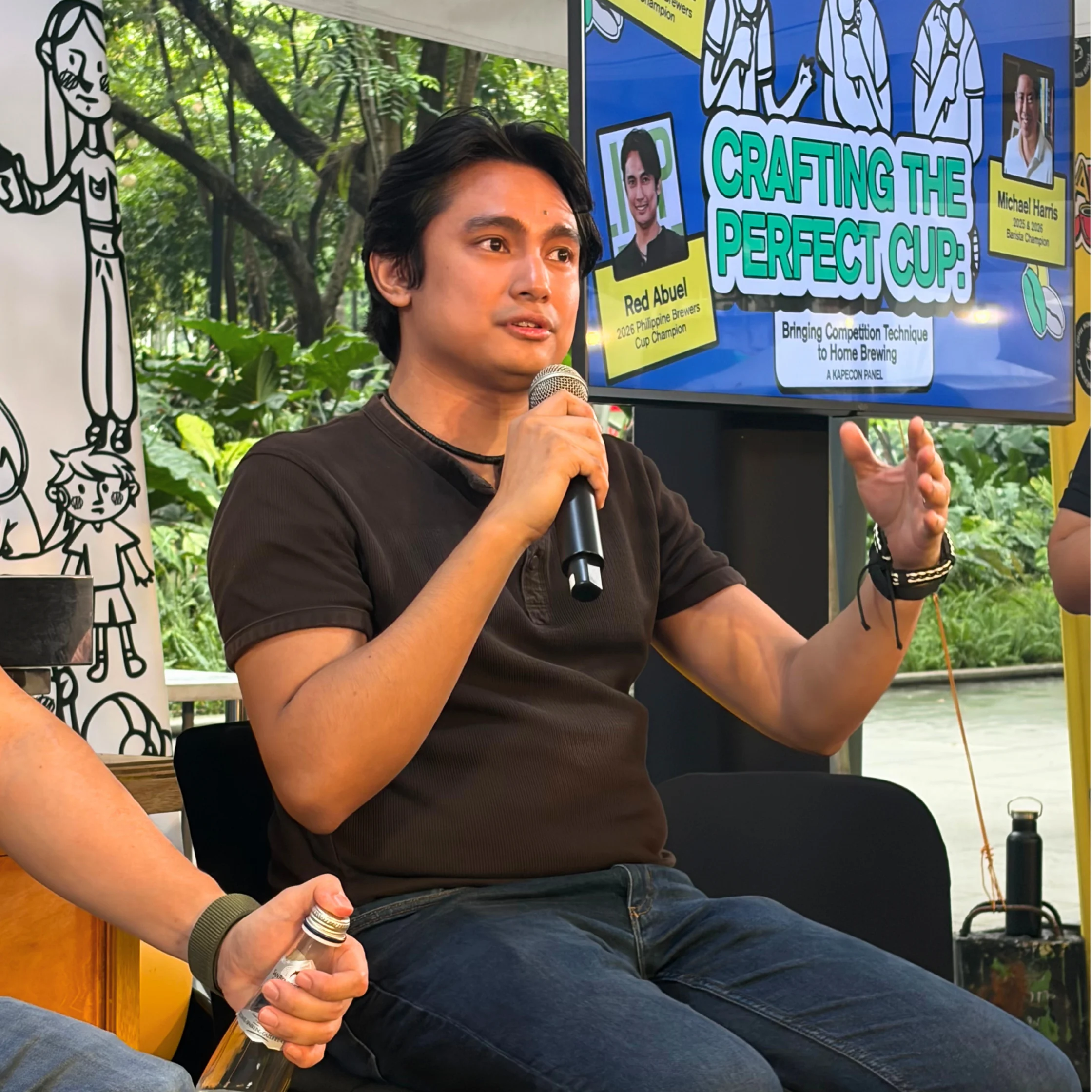 A seated man speaks into a microphone beside a screen reading “Crafting the Perfect Cup,” sharing a coffee presentation in a leafy outdoor setting.
