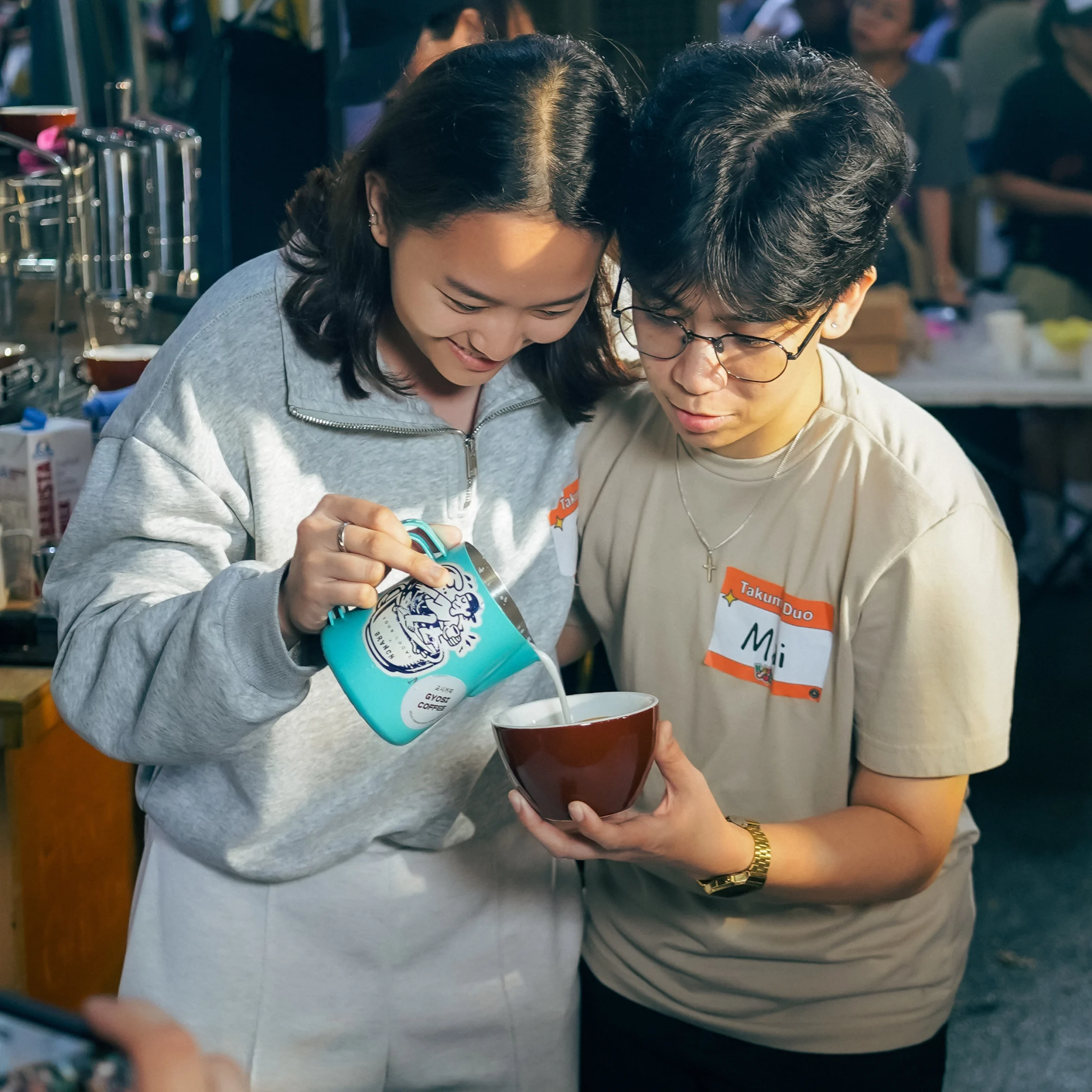 Two café workers smile as a woman pours milk from a turquoise pitcher into a cup held by her coworker, sharing a warm, focused moment.