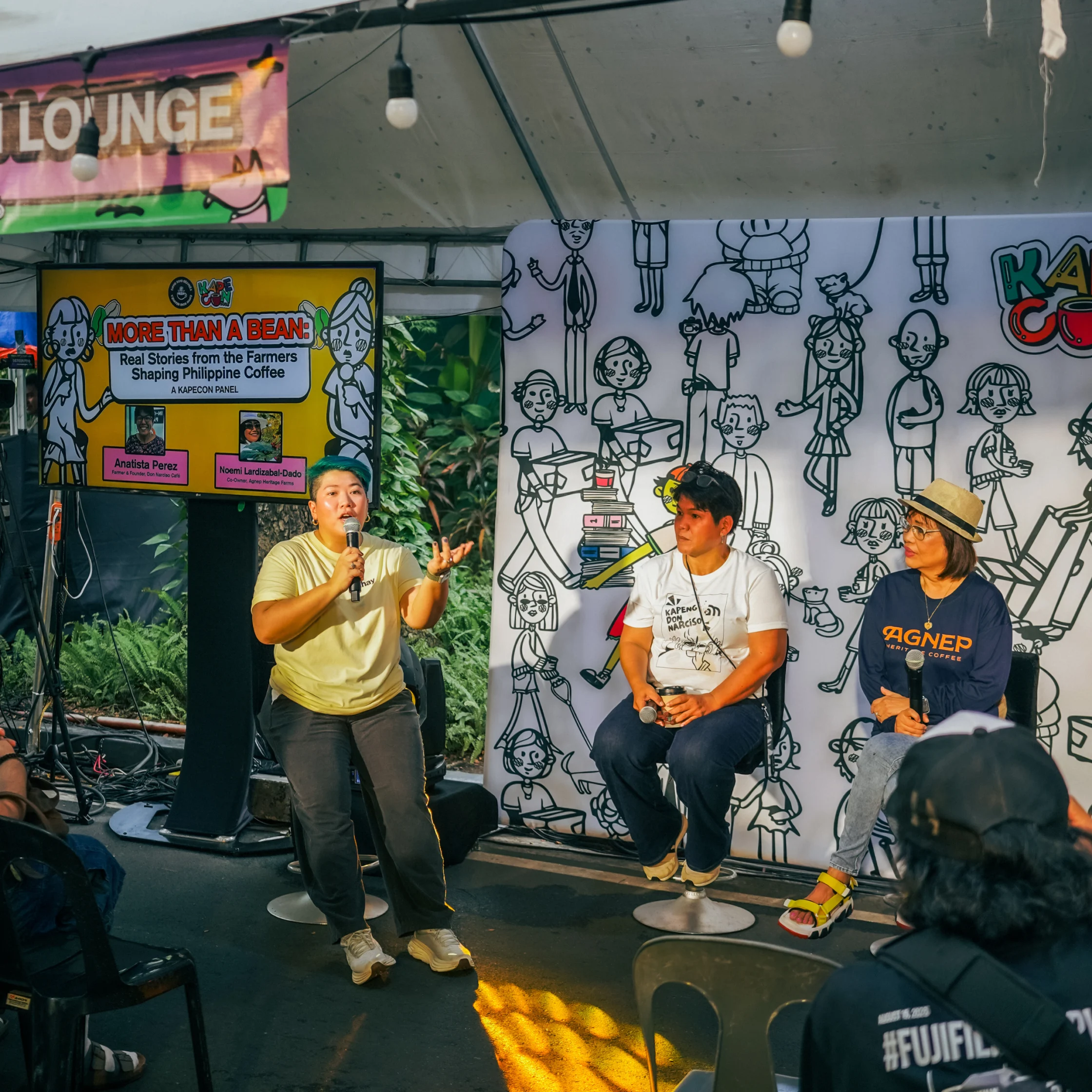 At a community event, a woman speaks into a microphone beside two seated panelists, with a coffee-farm poster and illustrated backdrop behind them.