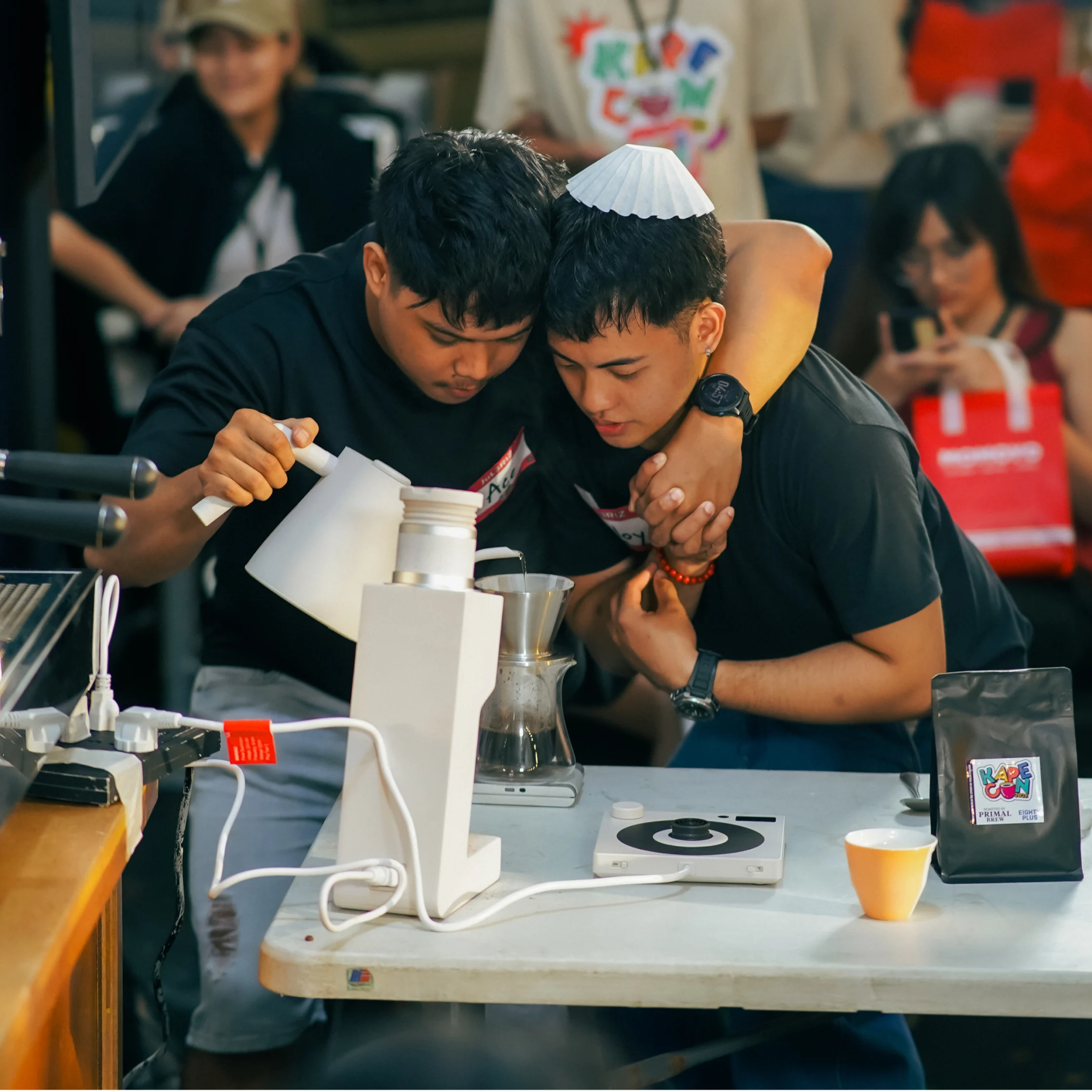 Two young men lean together over a tabletop science experiment, carefully pouring liquid into a glass vessel beside a small burner as onlookers watch; focused, collaborative mood.