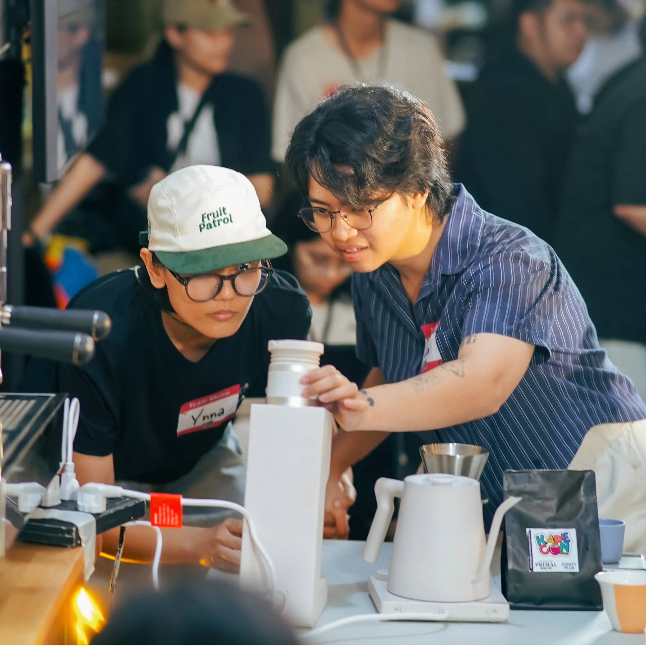Two young adults collaborate at a coffee-making station, carefully pouring into a cup among kettles and brewing tools; focused, friendly energy fills the busy event.