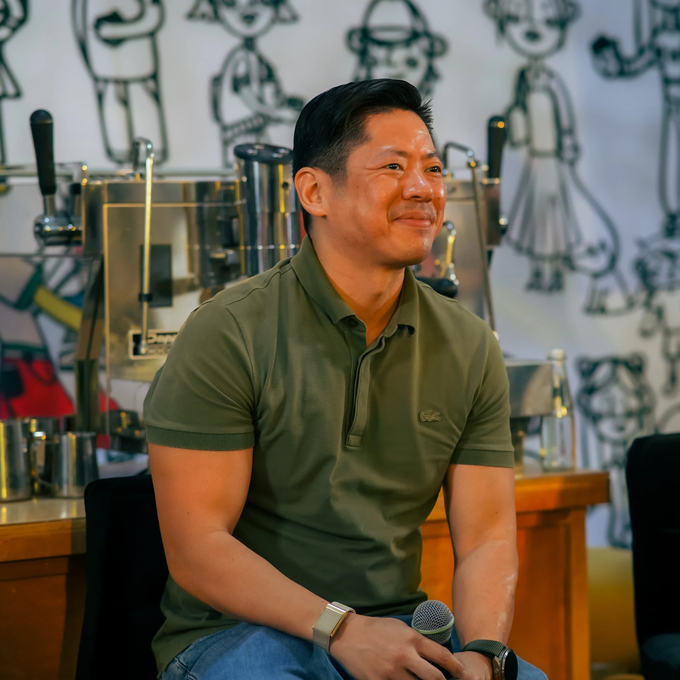 A smiling man in a green polo sits holding a microphone in a workshop, surrounded by metal equipment and playful character sketches.