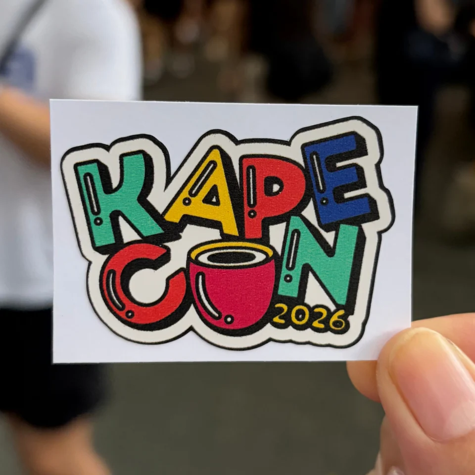 A hand holds a colorful “KAPE CON 2026” sticker featuring bold graffiti-style letters and a red coffee cup, with a blurred event crowd behind it.