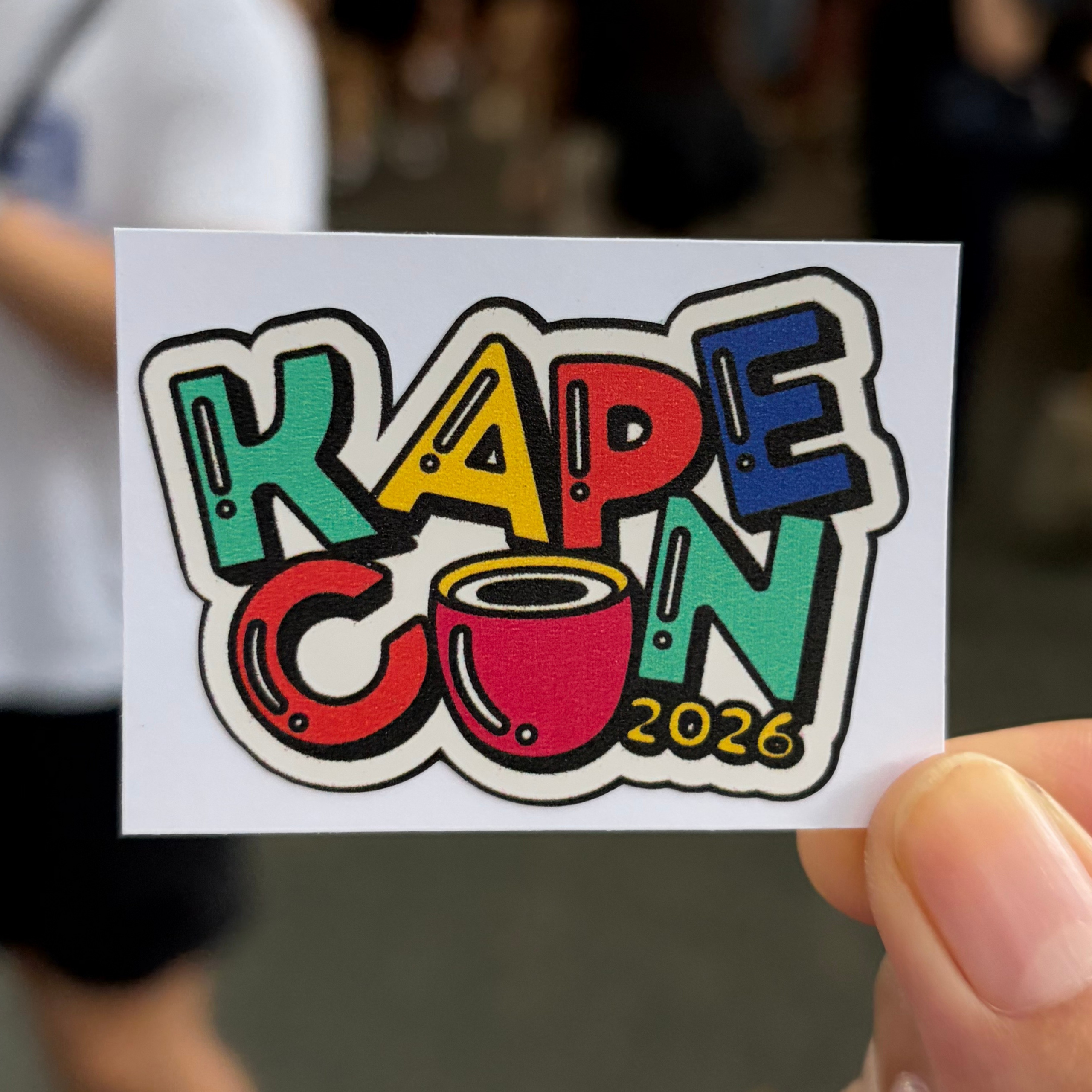 A hand holds a colorful “KAPE CON 2026” sticker featuring bold graffiti-style letters and a red coffee cup, with a blurred event crowd behind it.