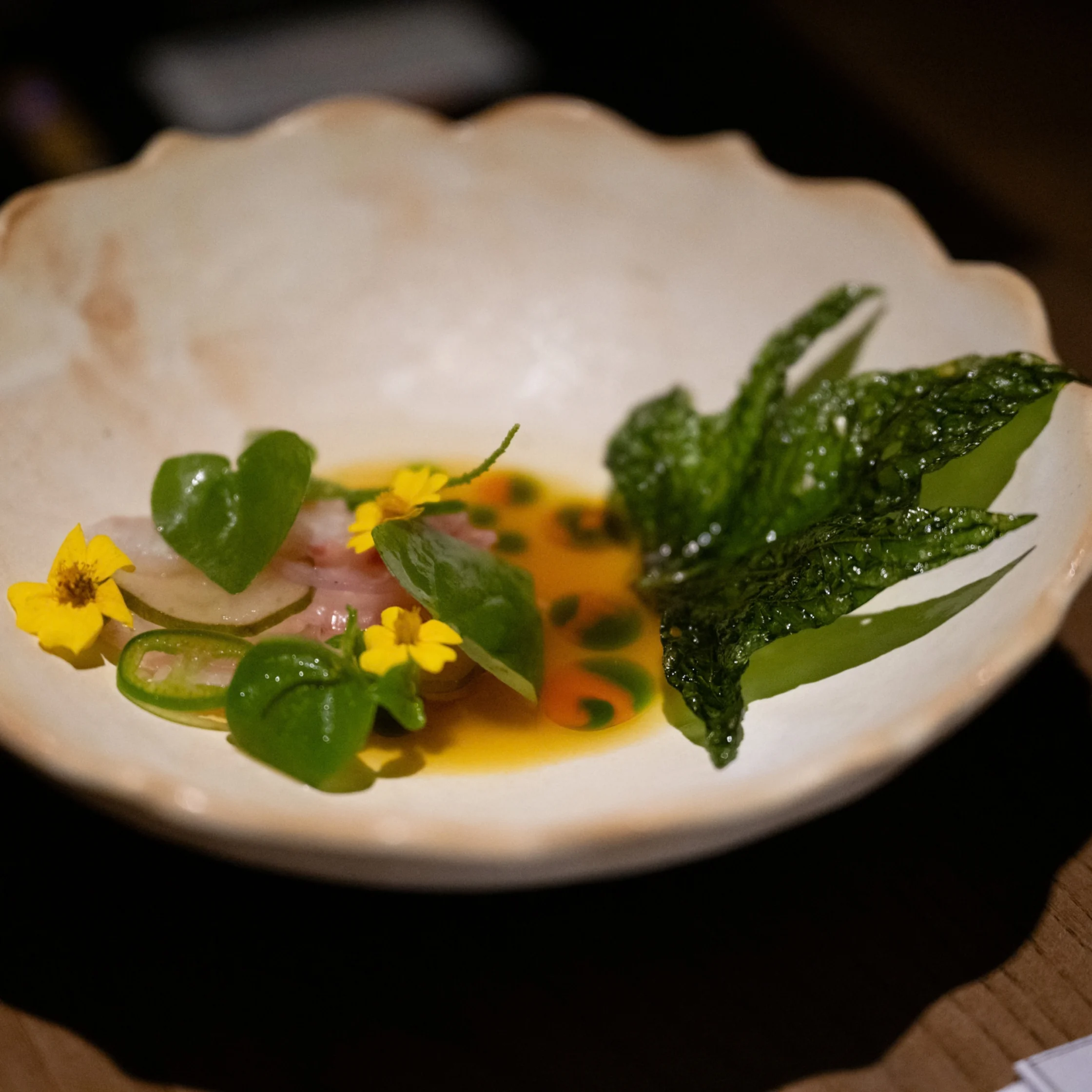 A refined plated dish of glossy green leaves, tiny yellow flowers, and fresh herbs beside amber sauce, arranged in a scalloped white bowl against a dark table.