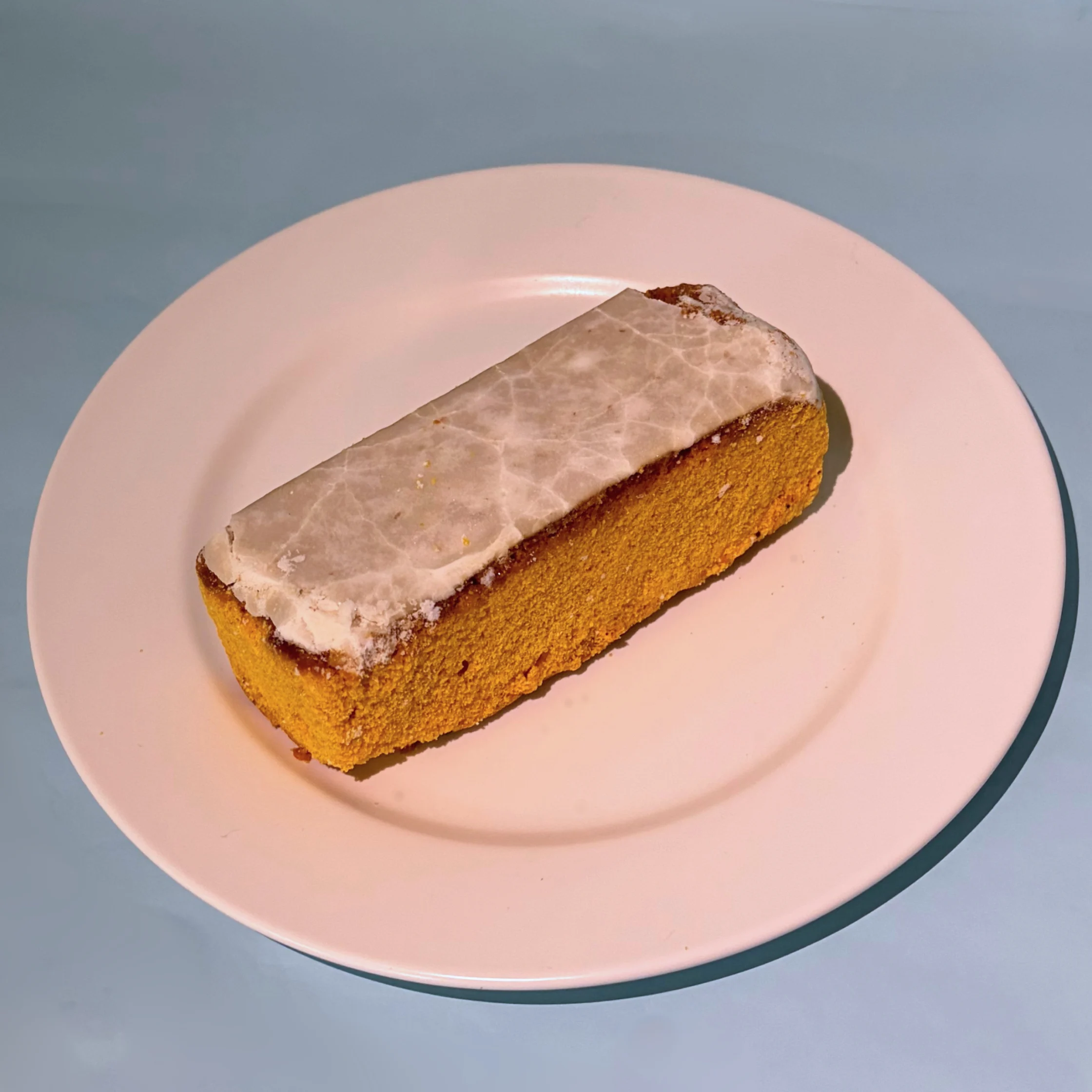 A golden-brown loaf cake topped with a thin layer of white icing sits alone on a pale pink plate against a cool blue-gray background.