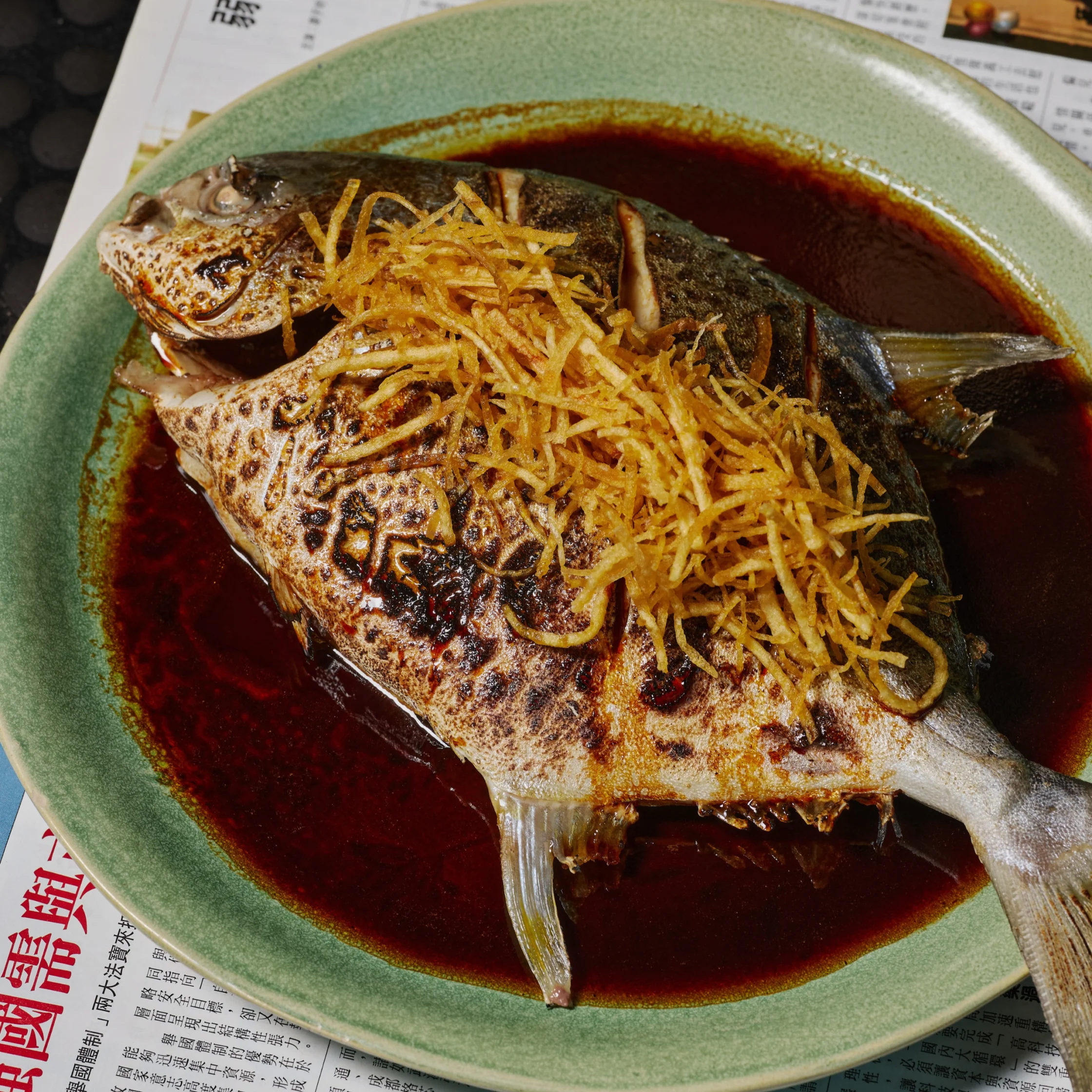 A whole grilled fish topped with crispy shredded ginger, served in a pool of dark sauce on a green-rimmed plate; a rich, appetizing dish.