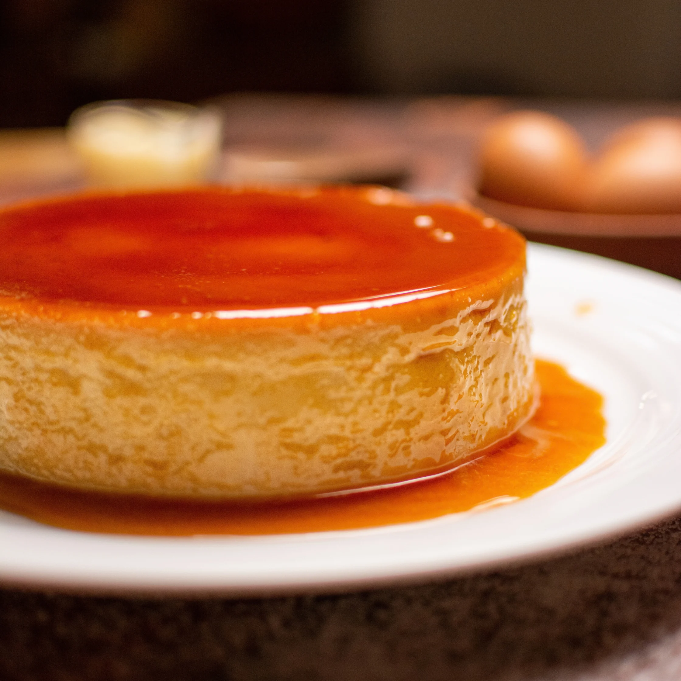A golden caramel flan sits on a white plate, its glossy amber topping and sauce gleaming in warm light against a softly blurred kitchen backdrop.
