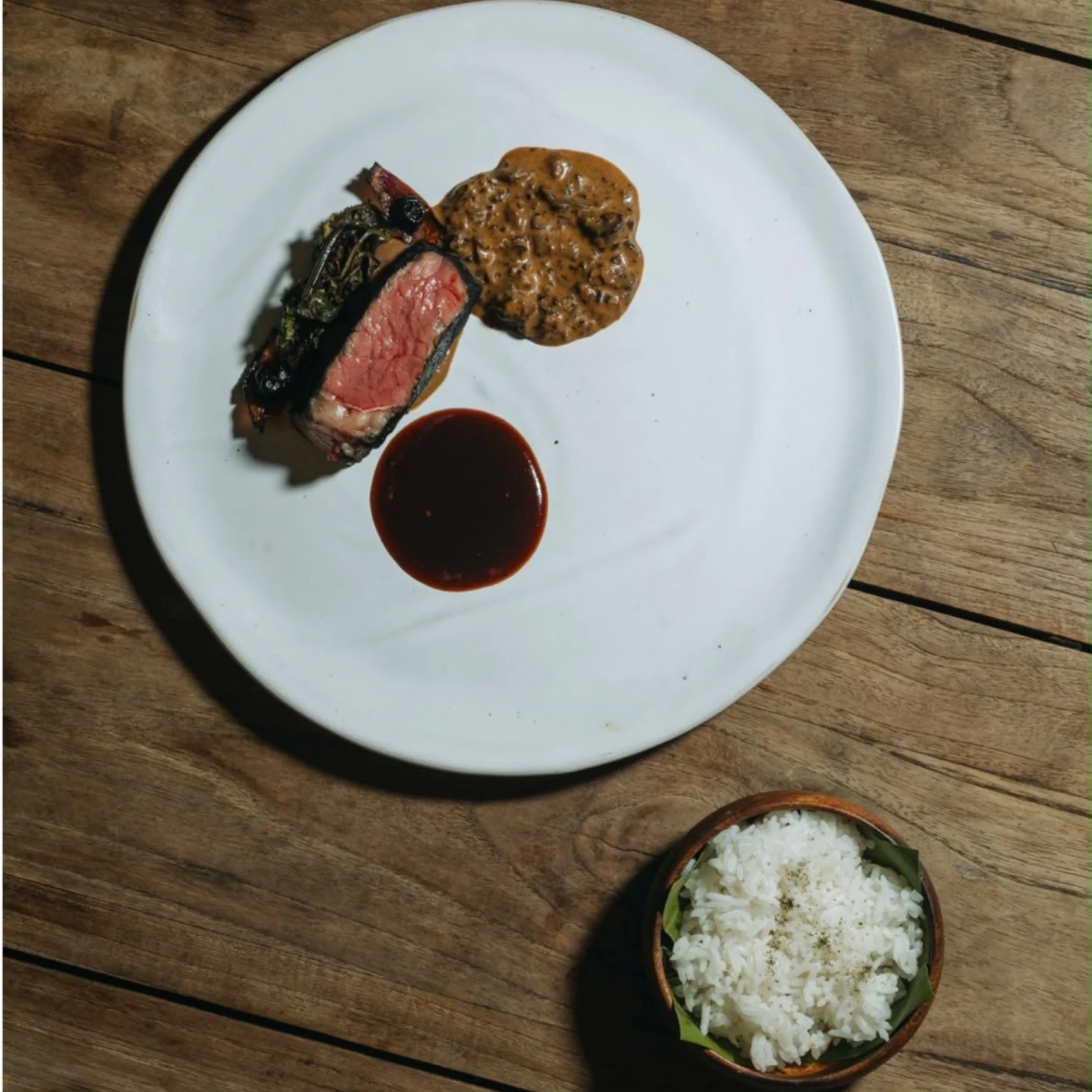 A white plate holds sliced medium-rare steak, creamy mushroom sauce, and a pool of red sauce beside a bowl of rice on a rustic wooden table.