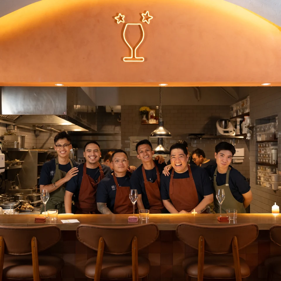 Six restaurant staff pose smiling behind the counter beneath a glowing orange arch and cocktail-glass logo, creating a warm, welcoming atmosphere.
