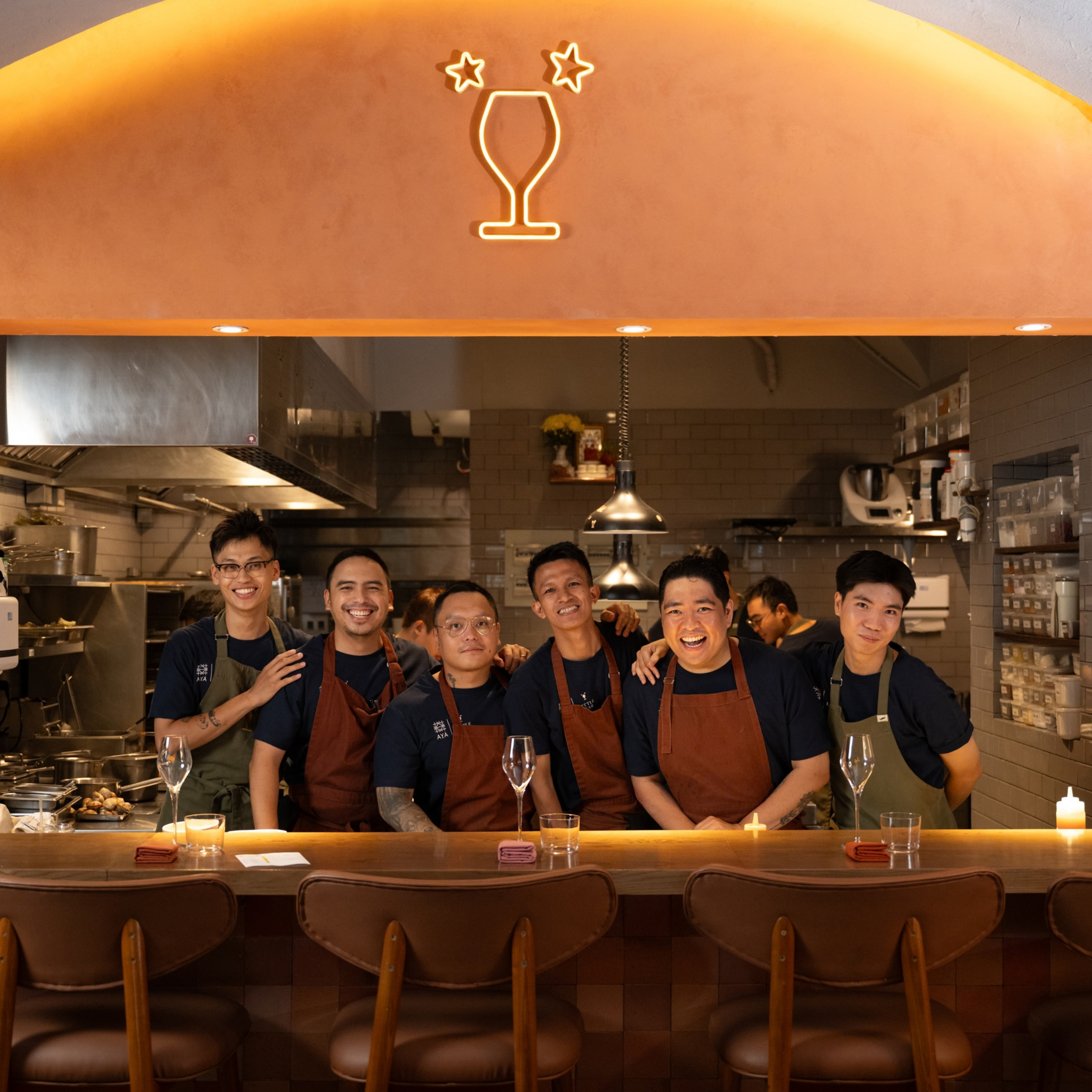 Six restaurant staff pose smiling behind the counter beneath a glowing orange arch and cocktail-glass logo, creating a warm, welcoming atmosphere.
