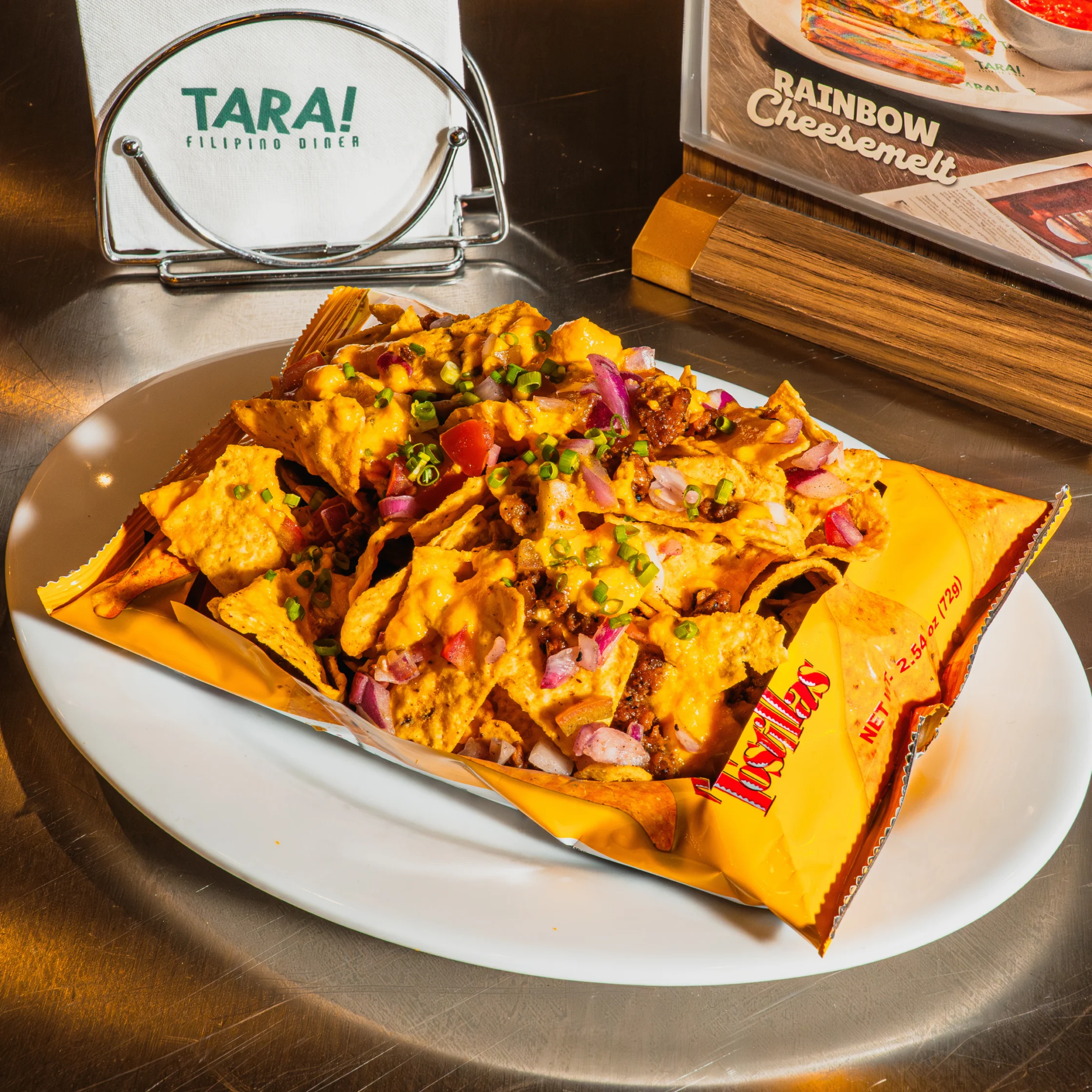 A vibrant Filipino-inspired nacho platter piled with chips, cheese, meat, tomatoes, and onions sits on a white plate beside a TARA! Filipino Diner sign.