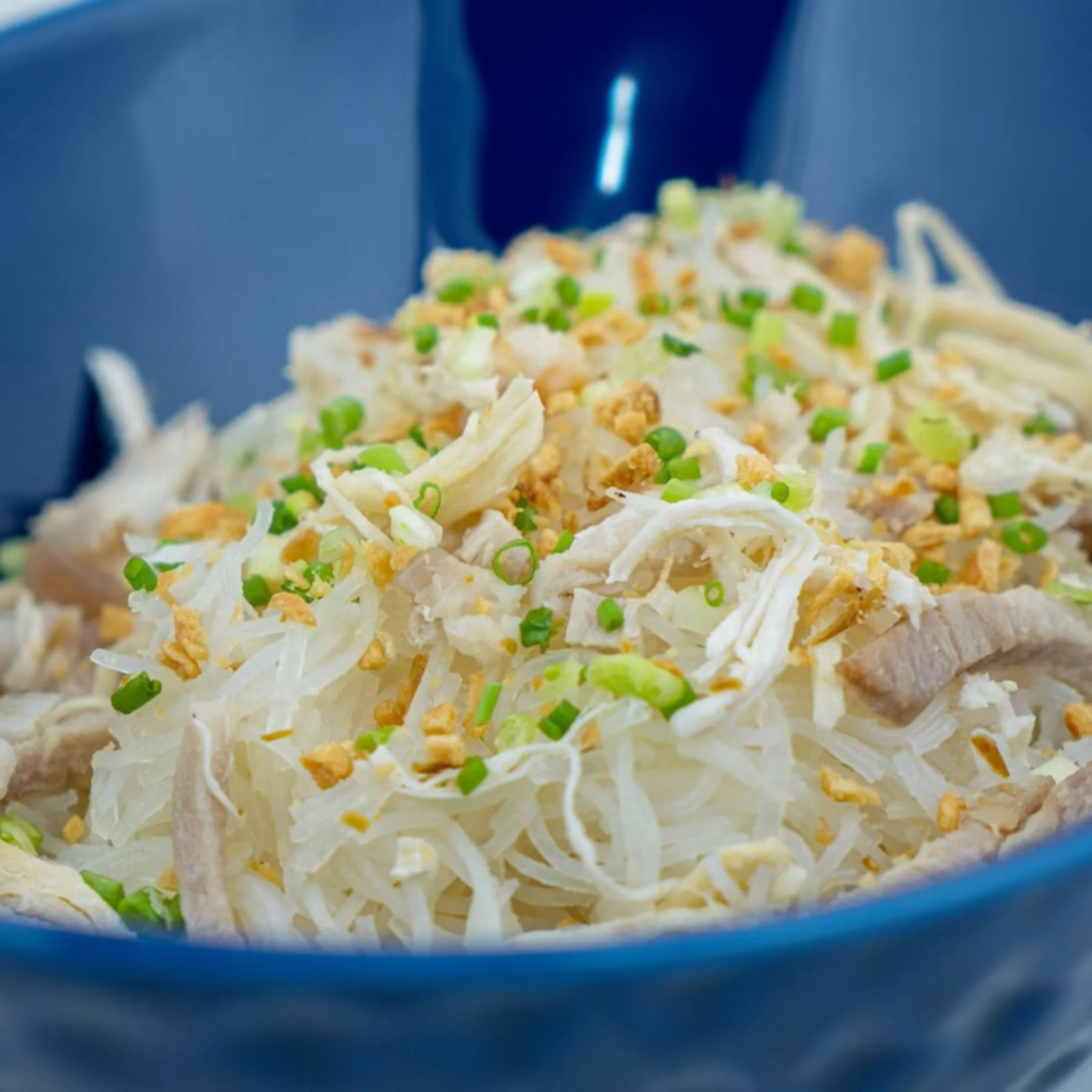 A blue bowl of shredded chicken and thin white noodles, topped with chopped scallions and crispy golden bits; a fresh, savory meal.