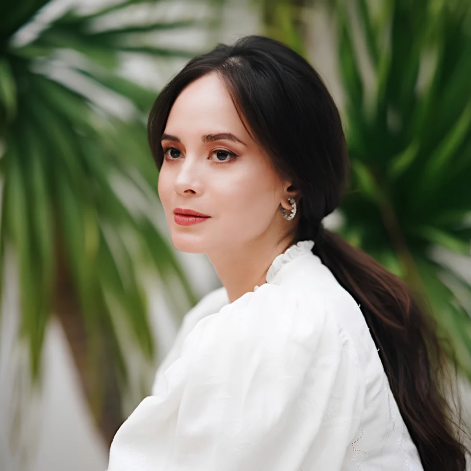 A poised woman with dark hair in a low ponytail wears a white blouse and silver earrings, gazing calmly amid lush green tropical leaves.