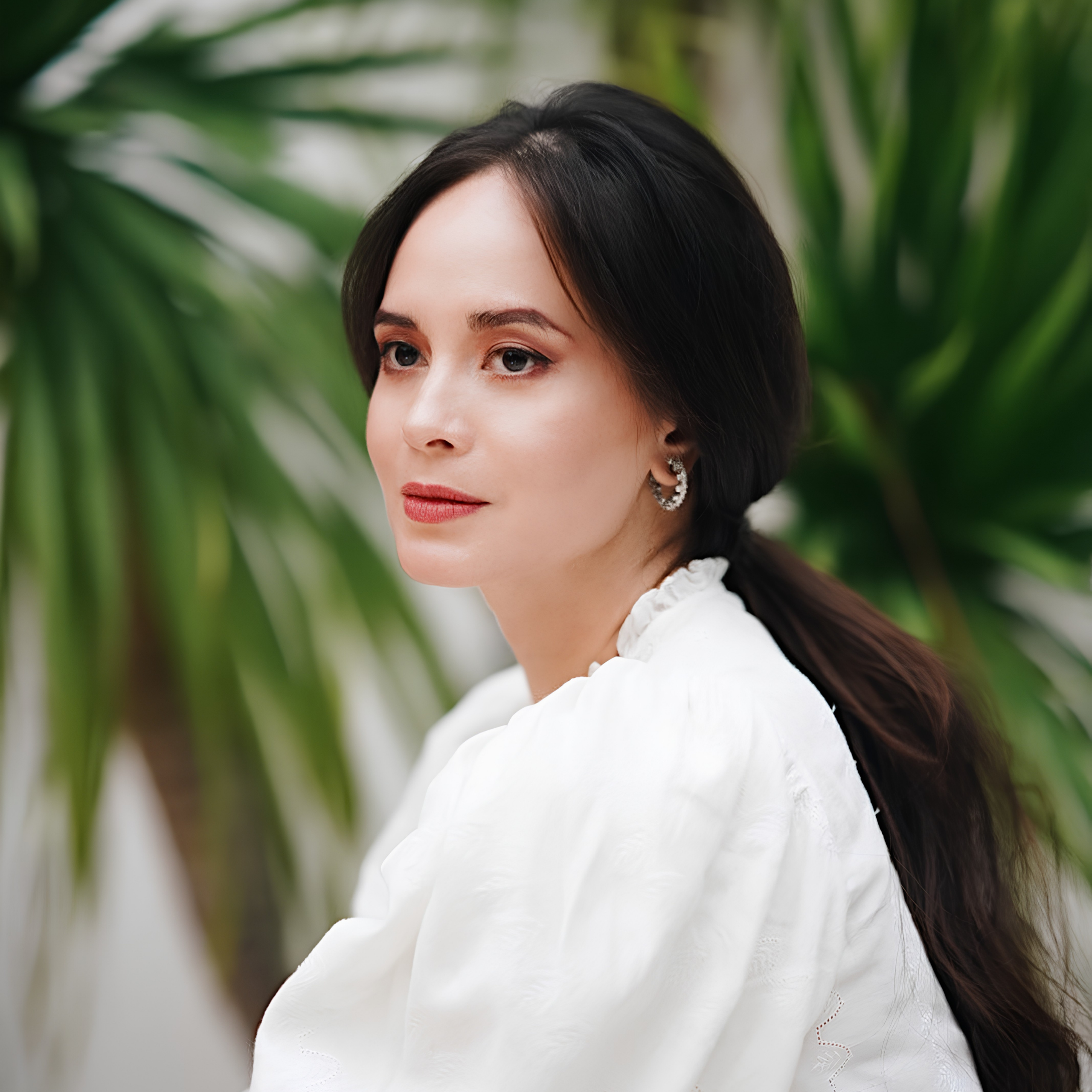 A poised woman with dark hair in a low ponytail wears a white blouse and silver earrings, gazing calmly amid lush green tropical leaves.