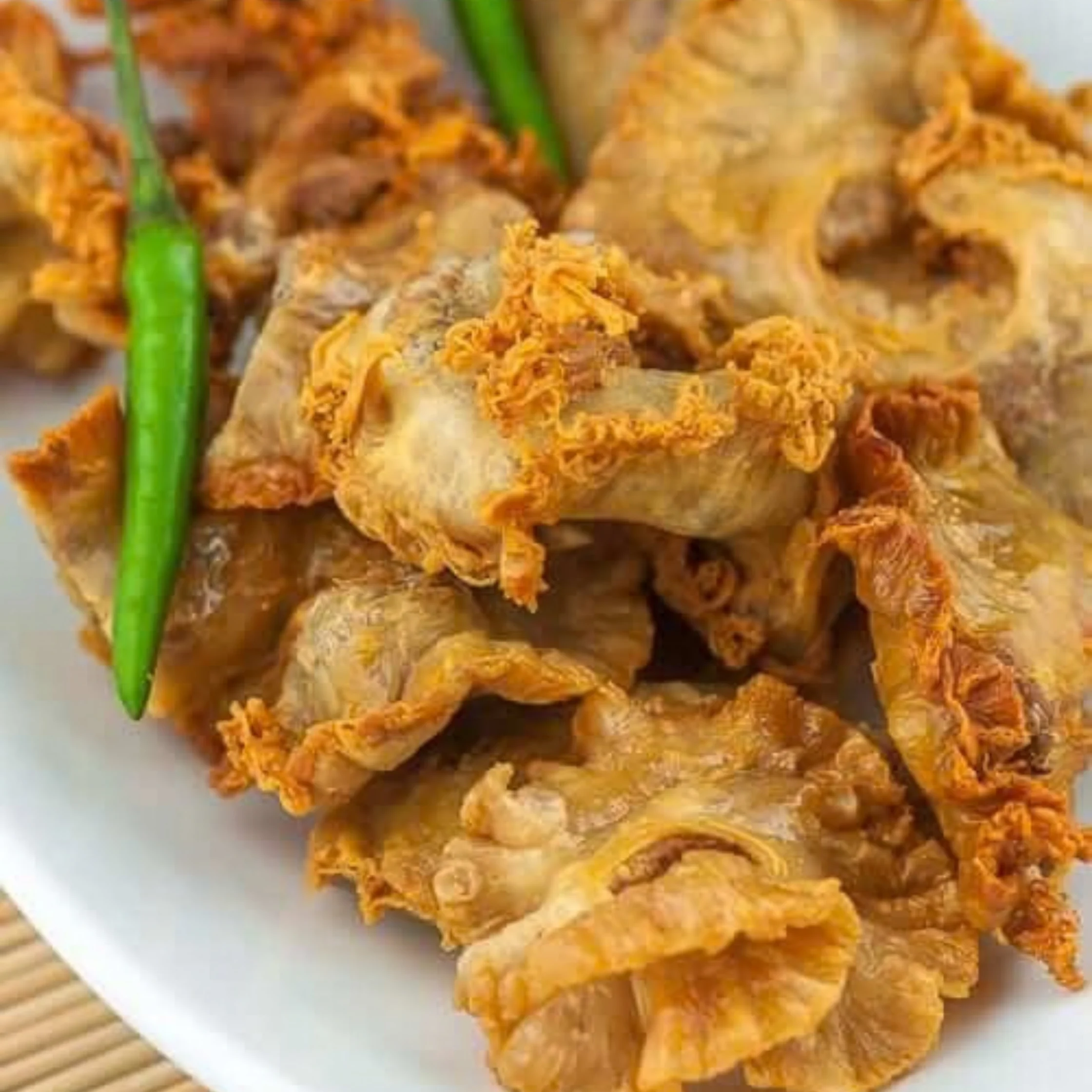 Golden, crispy fried meat piled on a white plate beside fresh green chilies, showing crunchy edges and tender pieces; the dish looks hearty and appetizing.