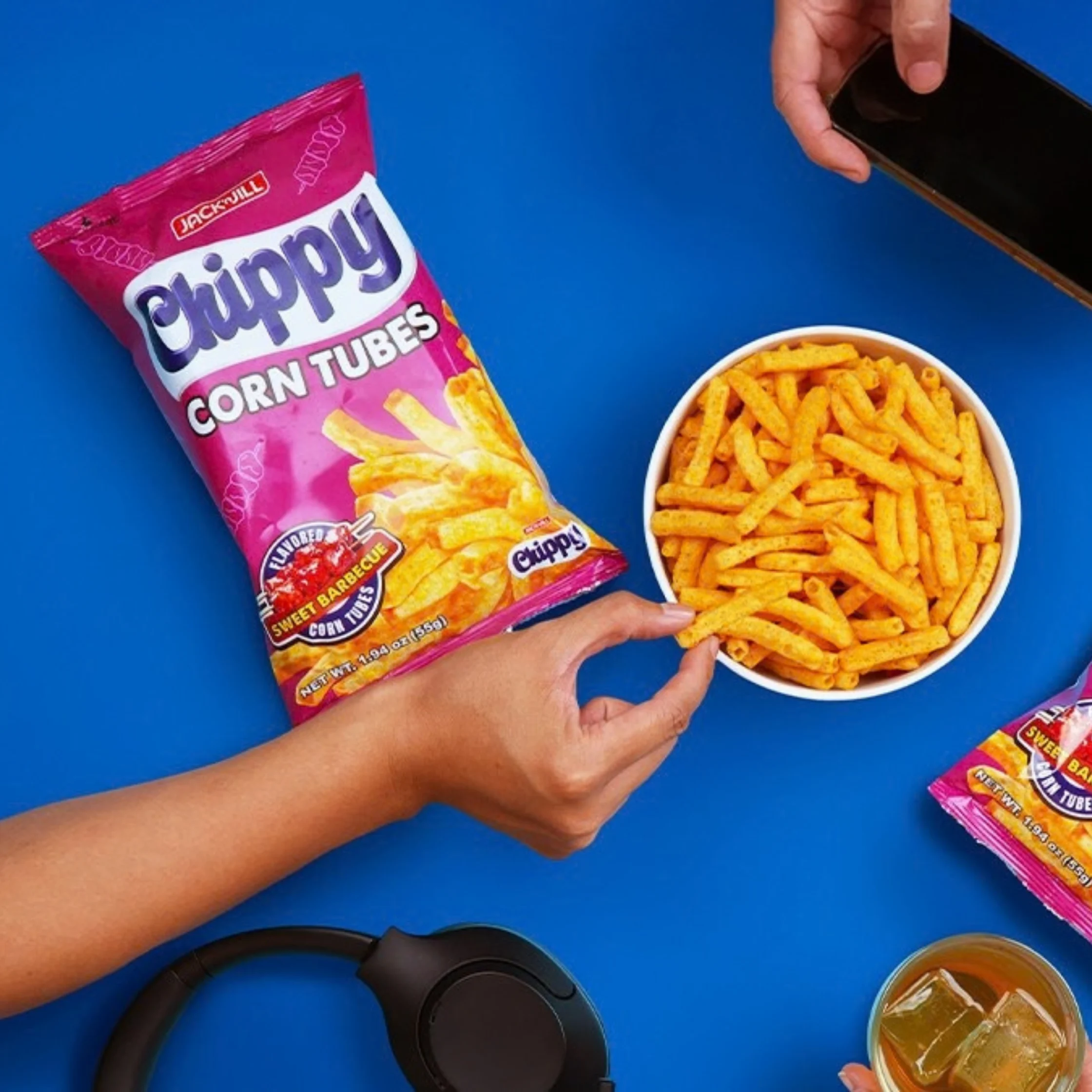 A hand reaches for orange corn tube snacks beside a bright pink Chippy bag on a blue surface, with headphones, a drink, and a phone nearby; playful snack-time scene.