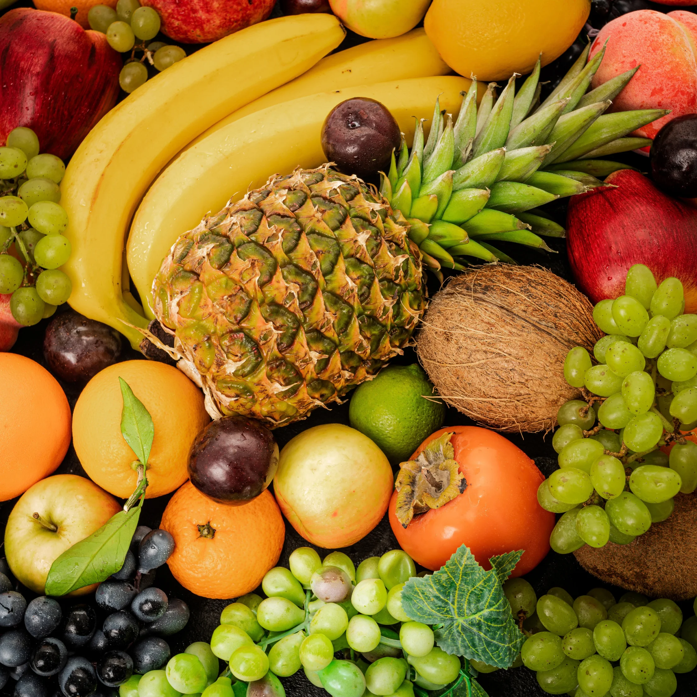 A colorful assortment of fresh tropical fruit, including pineapple, bananas, coconut, grapes, citrus, and apples, fills the frame with a vibrant, abundant feel.