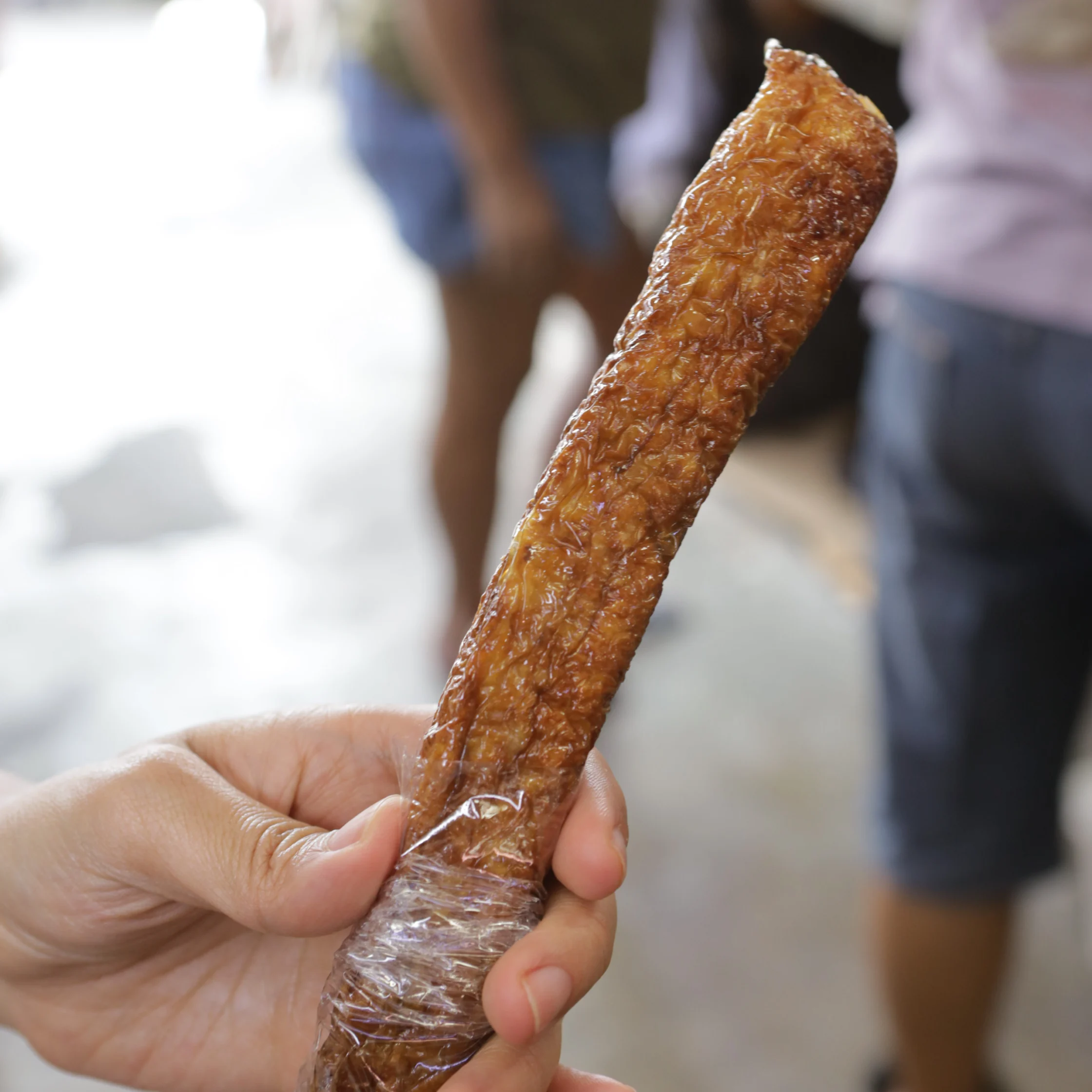 A hand holds a long, glossy brown meat snack wrapped in clear plastic, with blurred people in the background, suggesting a casual market setting.