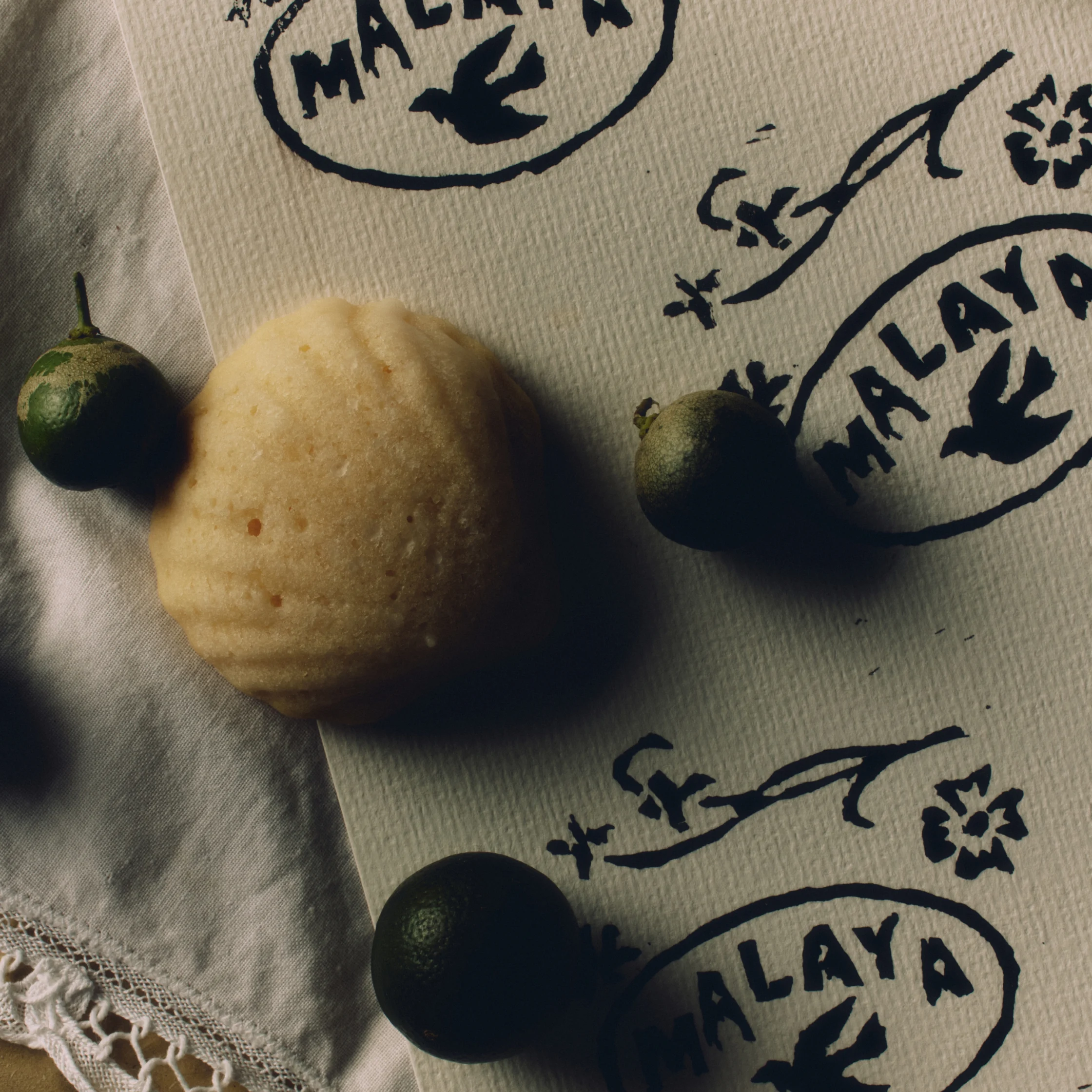 A pale, rough-skinned citrus fruit and three small dark green fruits rest on printed cloth in angled sunlight, creating a rustic, still-life mood.