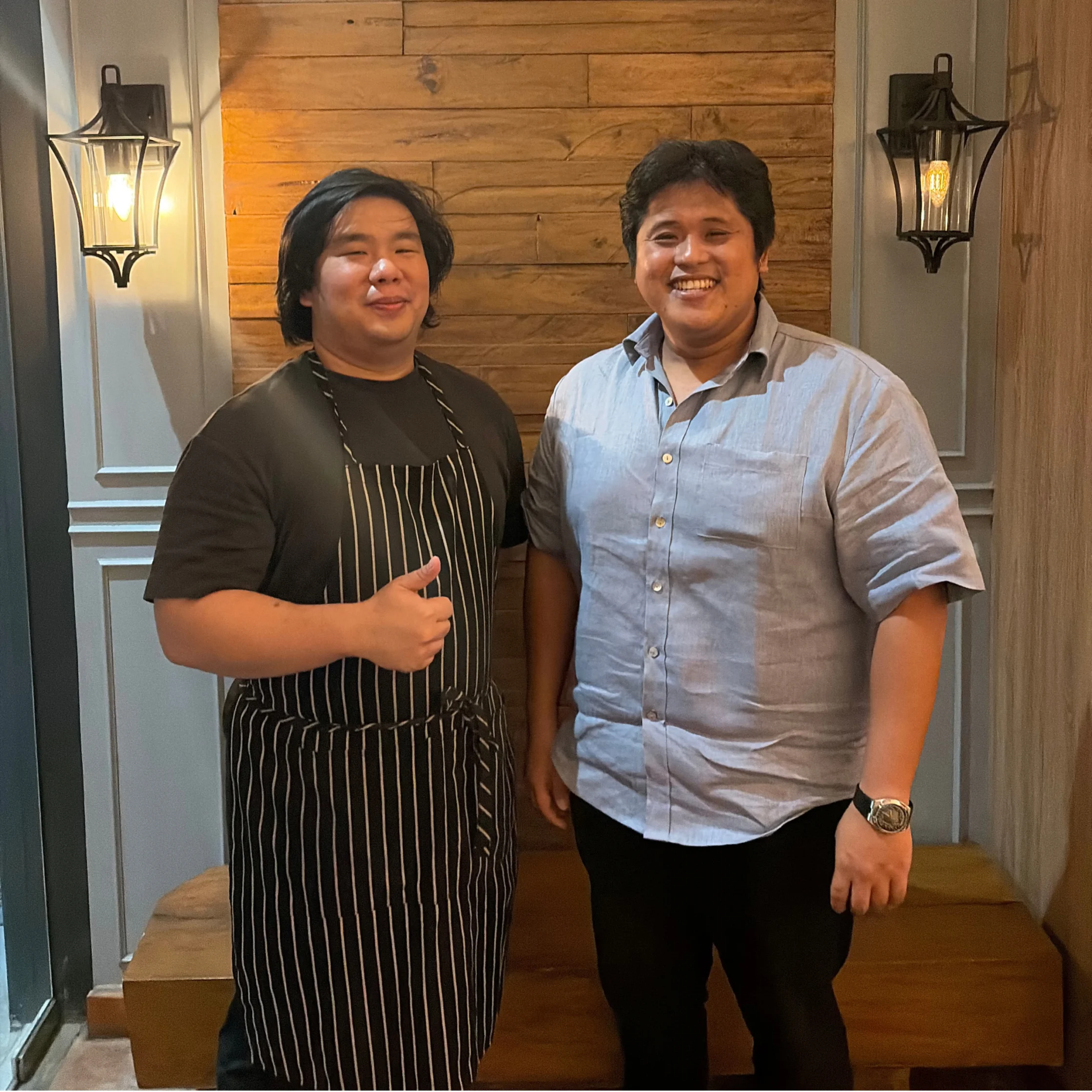 Two smiling men pose beside a wooden wall under warm lantern lights; one wears a striped apron and gives a thumbs-up, creating a friendly, welcoming mood.