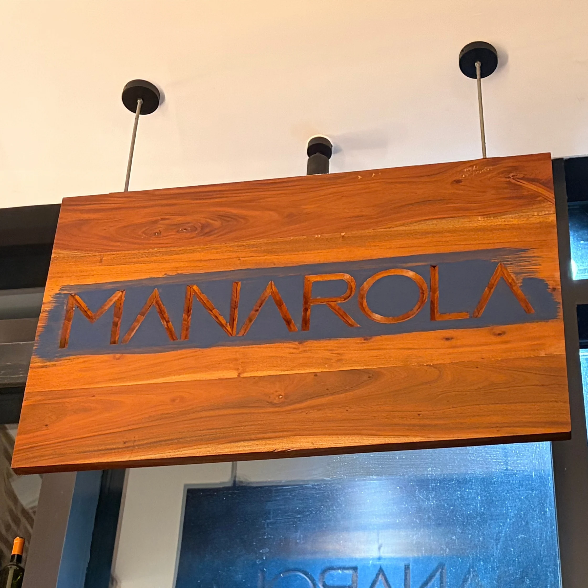 Warm wooden sign reading “MANAROLA” in large carved letters hangs above a glass doorway, evoking a welcoming, rustic Italian atmosphere.