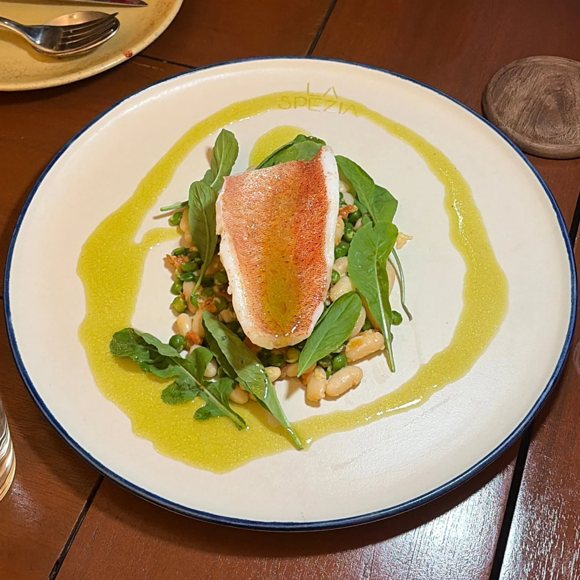 A seared pink fish fillet rests on greens, beans, and herbs, framed by a bright yellow sauce ring on a white plate; elegant restaurant presentation.