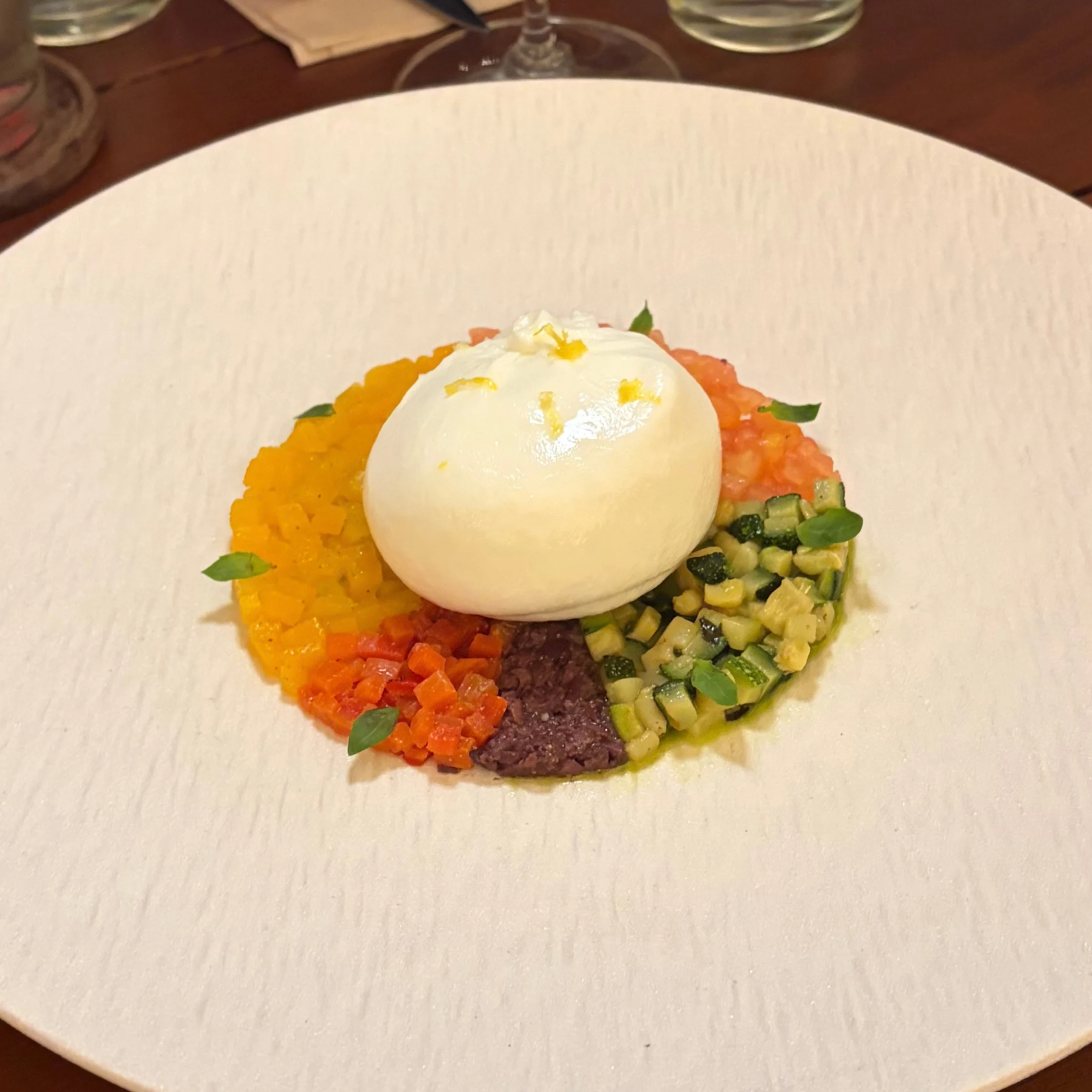 Creamy burrata sits over colorful diced vegetables and purée on a white plate, garnished with herbs; an elegant, vibrant restaurant dish.