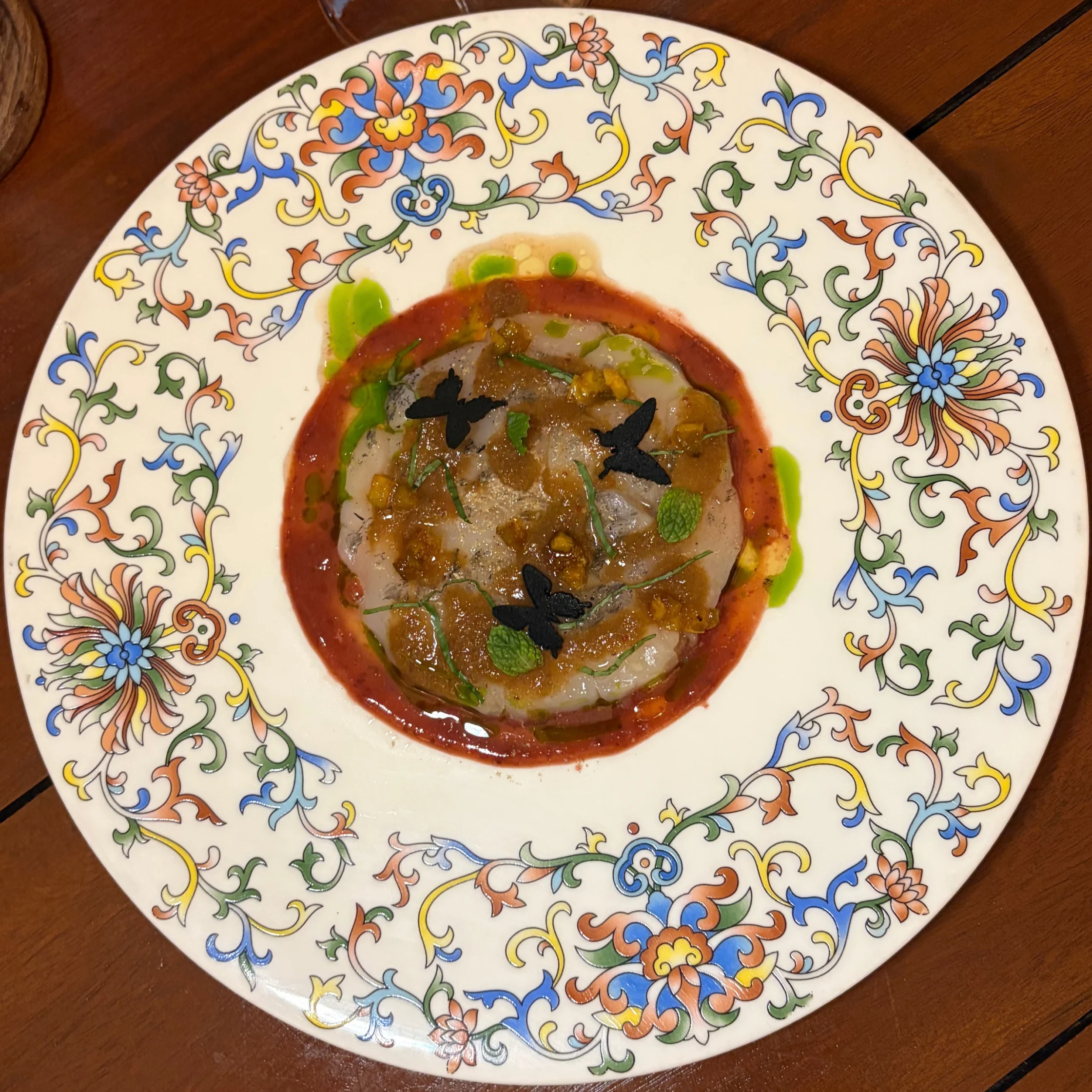 An elegant plated dish of layered vegetables in red sauce, topped with herbs and black olives, sits on an ornate floral plate against a wooden table.